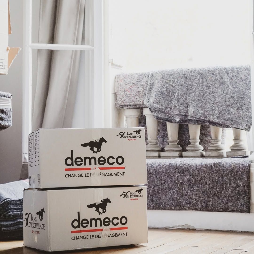 Deux cartons Demeco empilés à côté d’une fenêtre et d’une banquette grise rembourrée, dans une pièce lumineuse