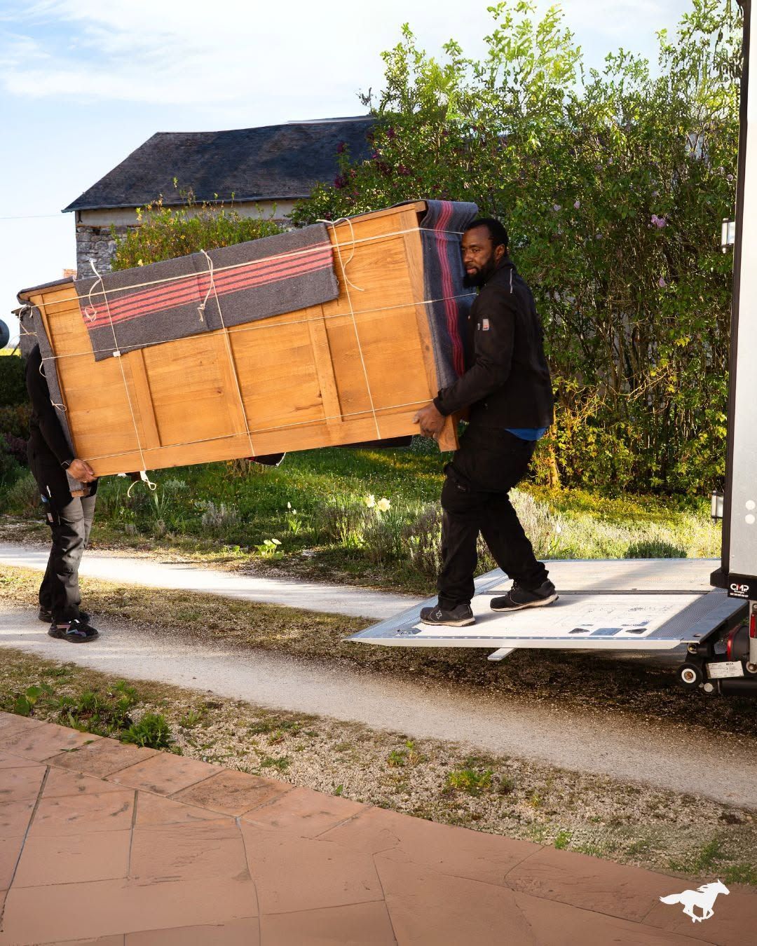 Deux personnes transportent une grande caisse en bois près d’un camion, dans une allée