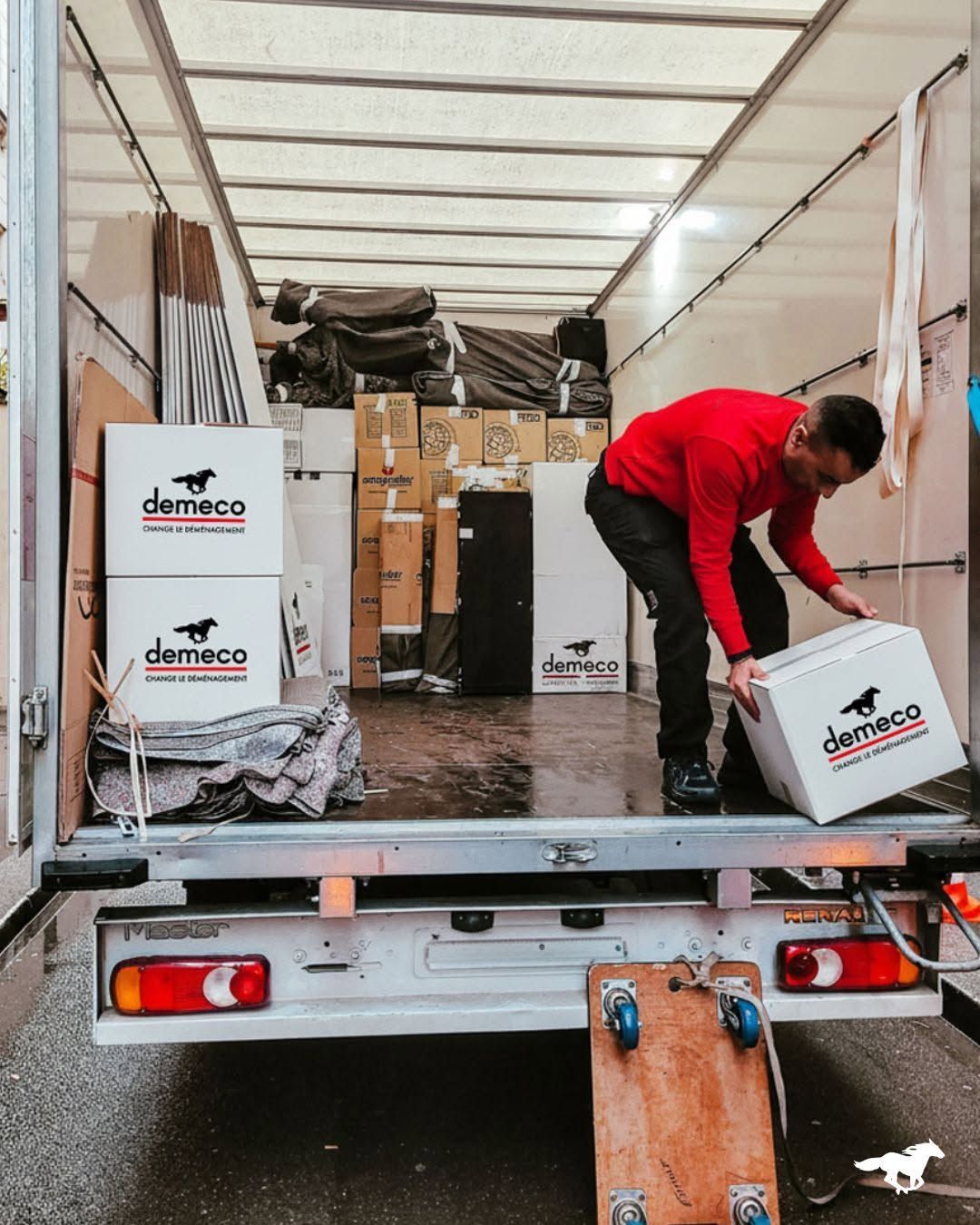 Un employé en veste rouge charge des cartons dans un camion de livraison.