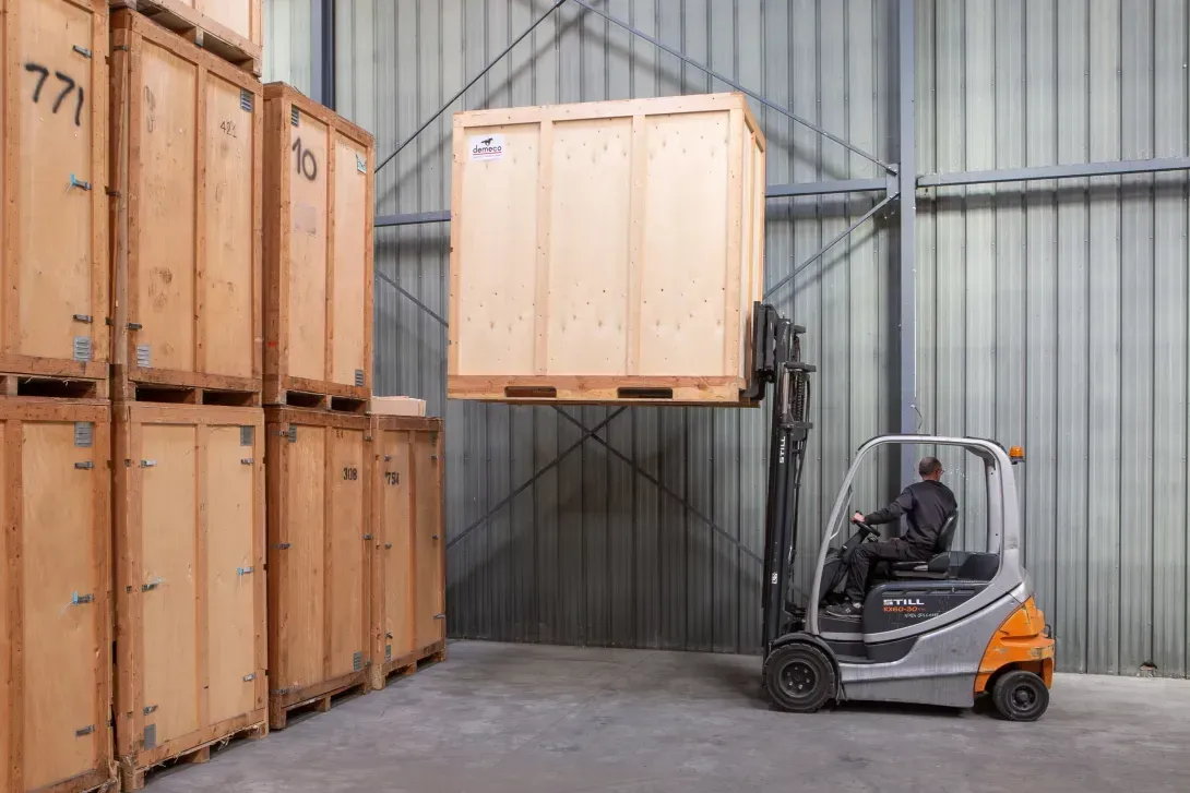 Un chariot élévateur soulève une caisse en bois dans un entrepôt, à côté de caisses empilées.