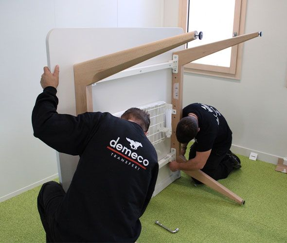 Deux ouvriers soulèvent et déplacent une armoire blanche dans une pièce lumineuse avec une moquette verte.