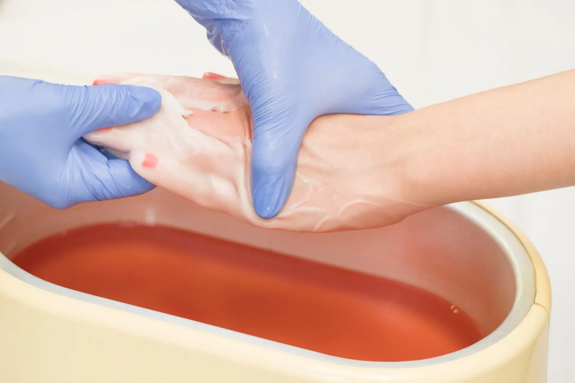 Manos con guantes azules sumergiendo un pie en un baño de cera tibia para un tratamiento de spa.