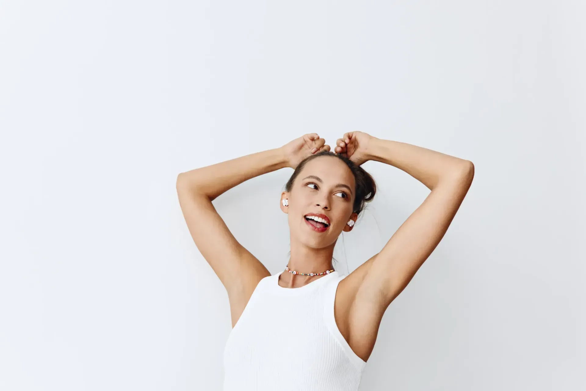 Mujer con top blanco, brazos levantados, cabello recogido, sonriendo, usando auriculares, sobre un fondo blanco.