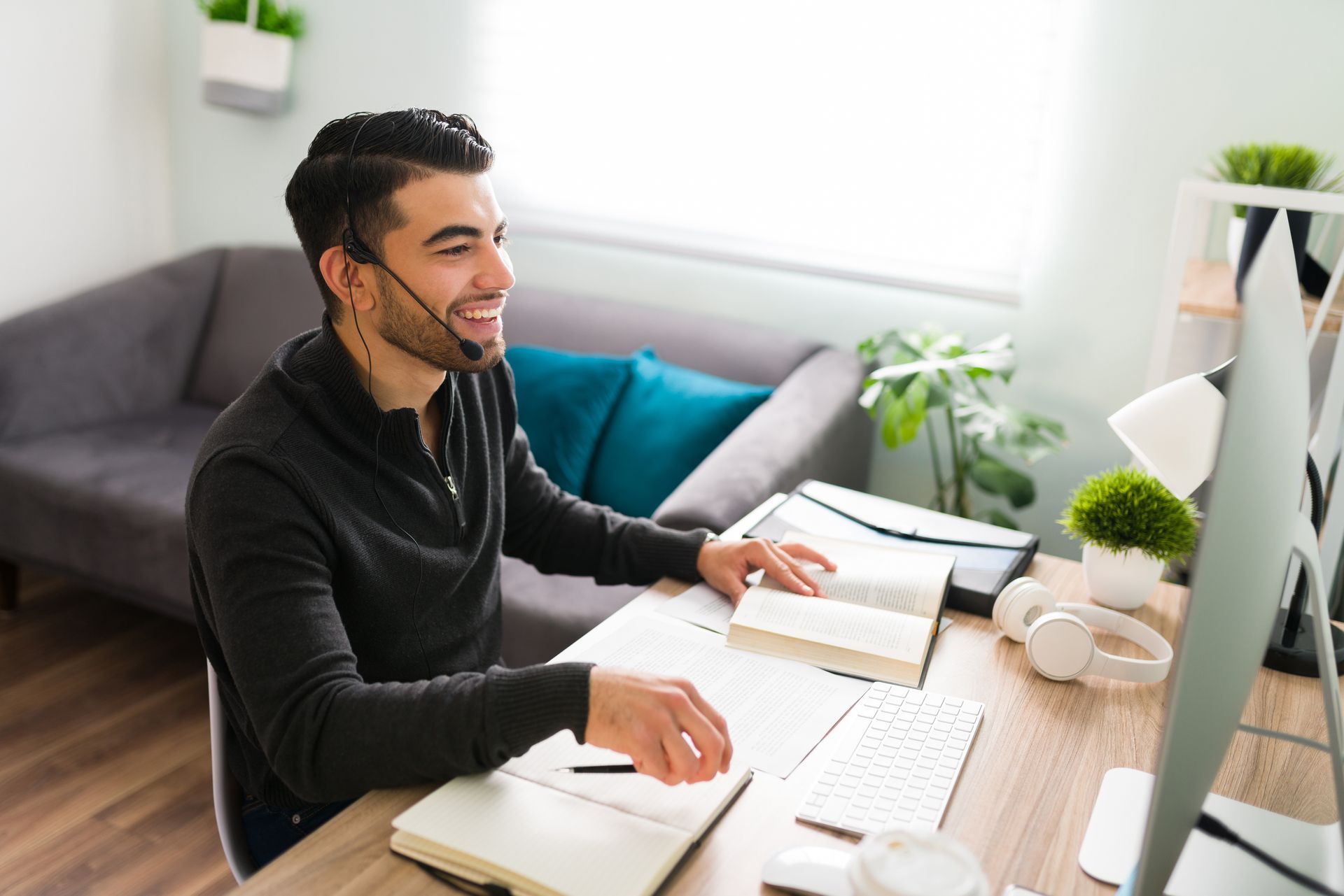 Une personne portant un casque audio sourit en travaillant à un bureau avec un ordinateur, un livre ouvert et un carnet dans un bureau à domicile lumineux.