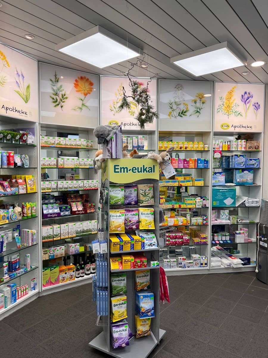 Innenraum einer Apotheke mit Produktregalen, beleuchtet durch Deckenlampen. Ausstellungsständer mit Gesundheitsprodukten in der Mitte.