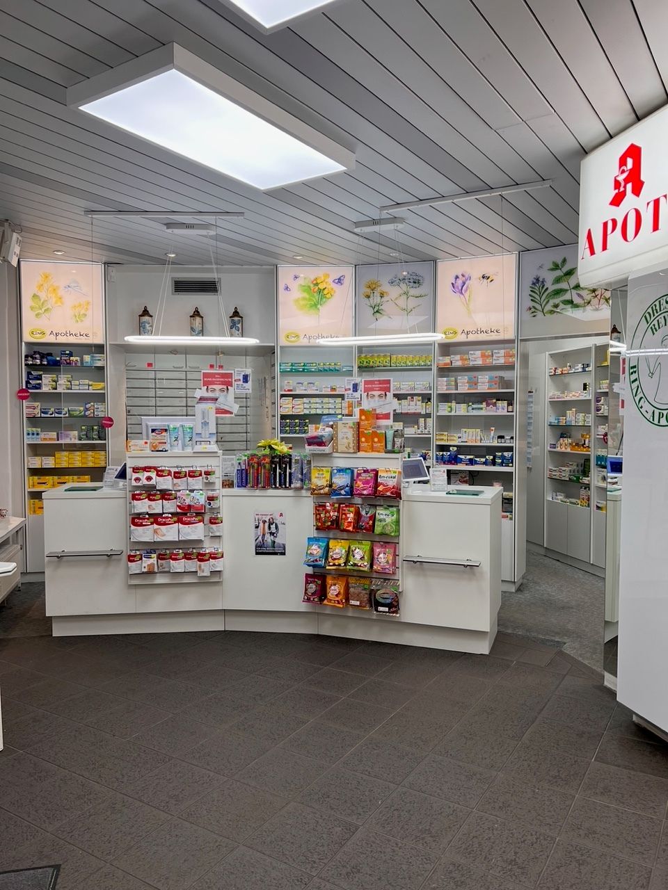 Innenraum einer Apotheke mit weißen Theken, Medikamentenregalen und einem roten Apothekersymbol.