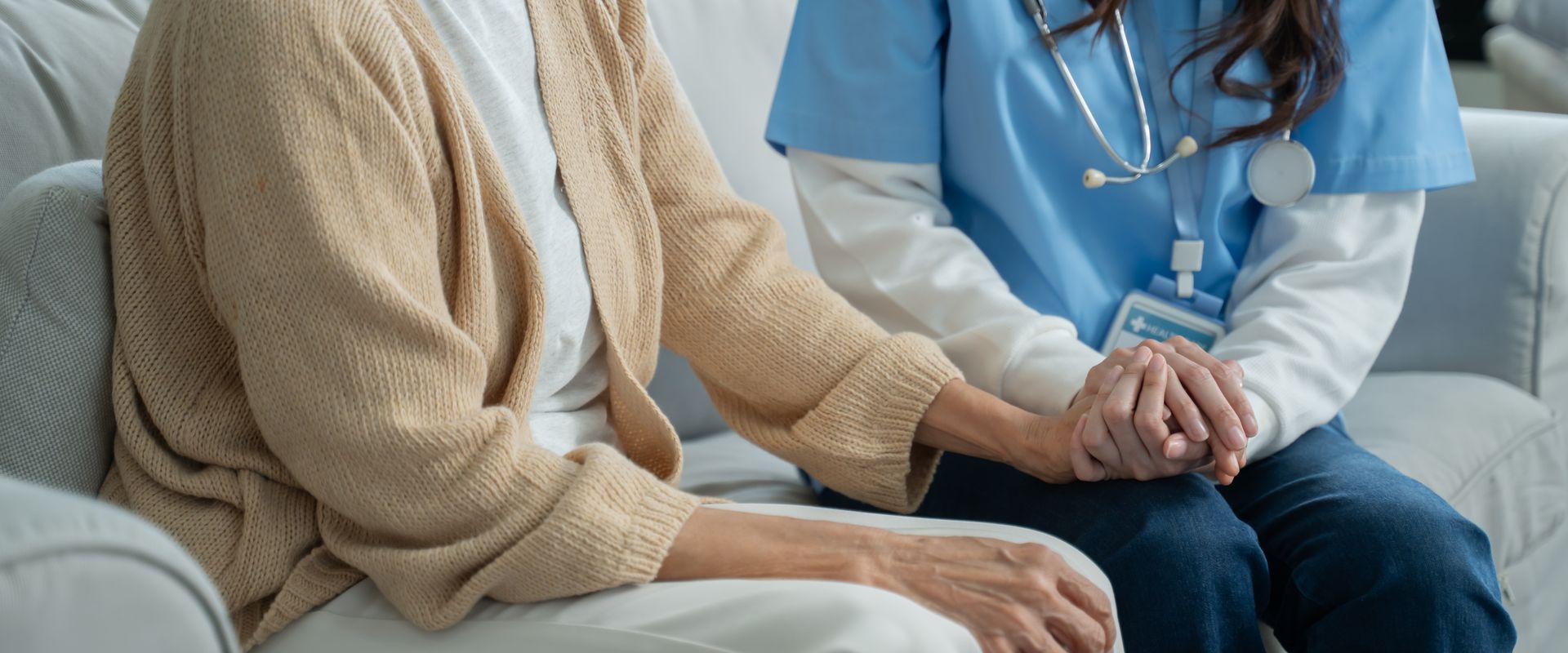 Una donna anziana e un'infermiera si tengono per mano su un divano. L'infermiera indossa il camice.
