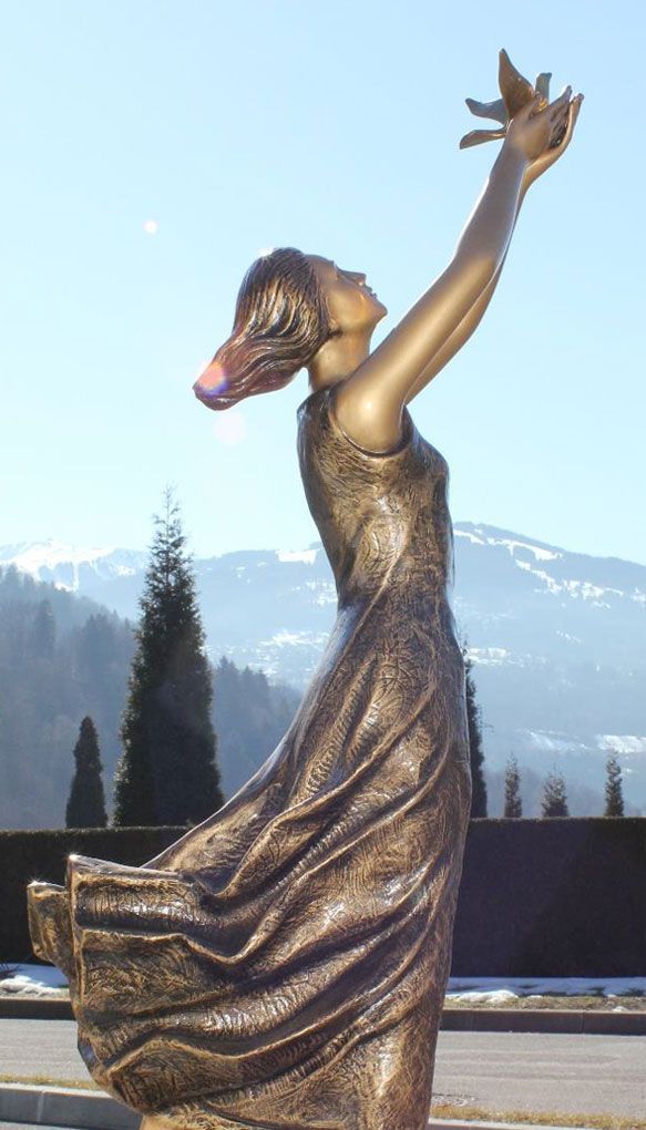 Statue en bronze d'une femme aux bras levés, tenant un oiseau, robe flottante. Montagnes et arbres en arrière-plan.
