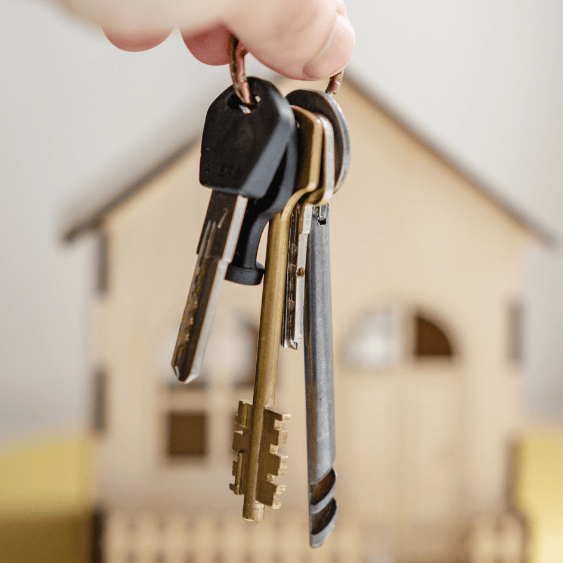 Mano sosteniendo un llavero con un objeto en forma de casa al fondo, simbolizando la propiedad de una vivienda.