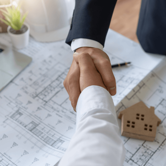 Dos personas estrechando la mano sobre unos planos arquitectónicos y una pequeña casa de madera.