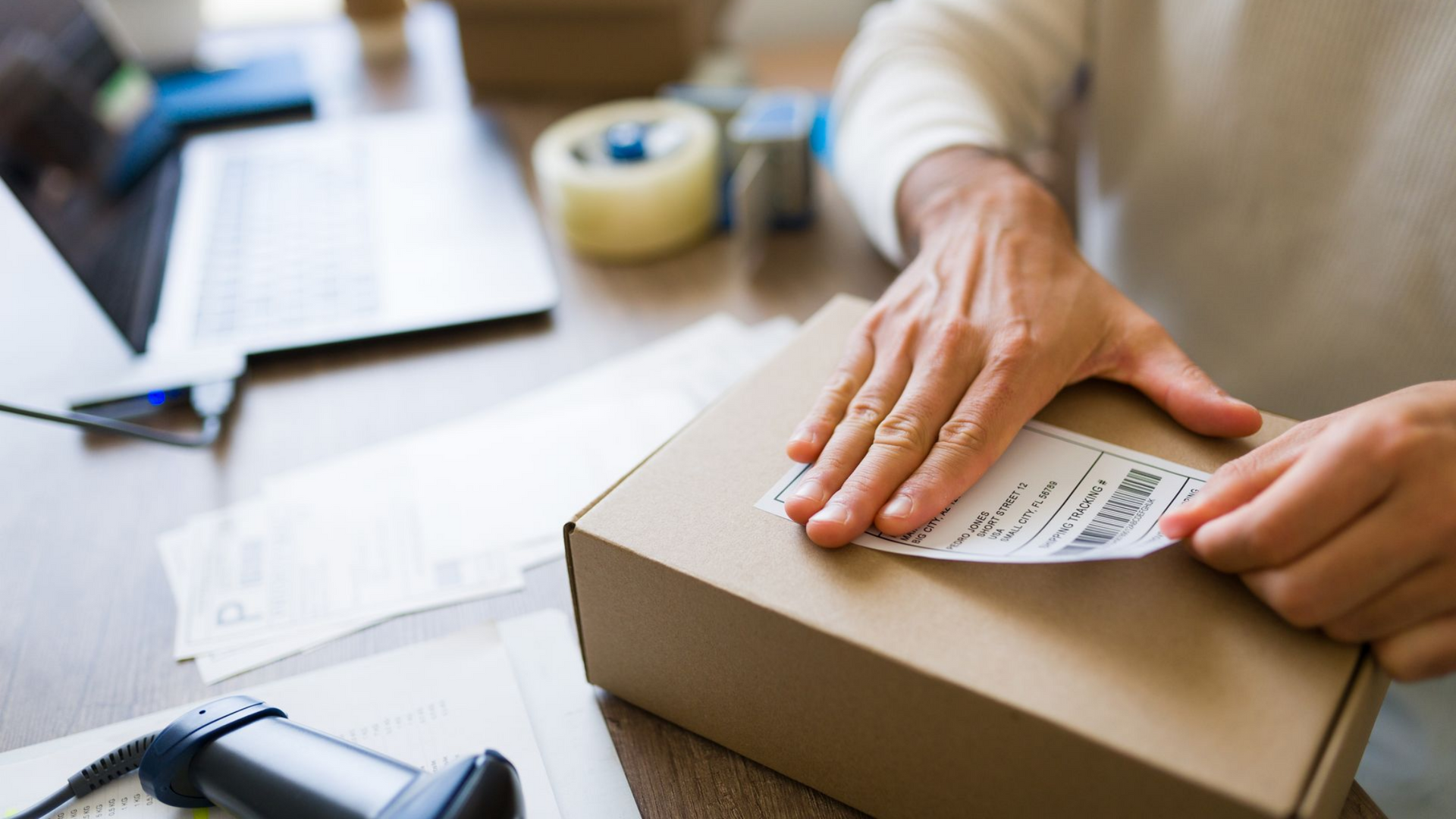 Une personne colle une étiquette d'expédition sur une boîte en carton, travaillant à un bureau avec un ordinateur portable et un lecteur de codes-barres.