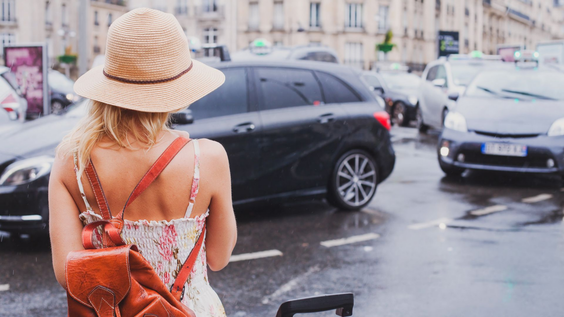 Femme avec un sac à dos et un chapeau regardant les voitures dans une rue mouillée de la ville.