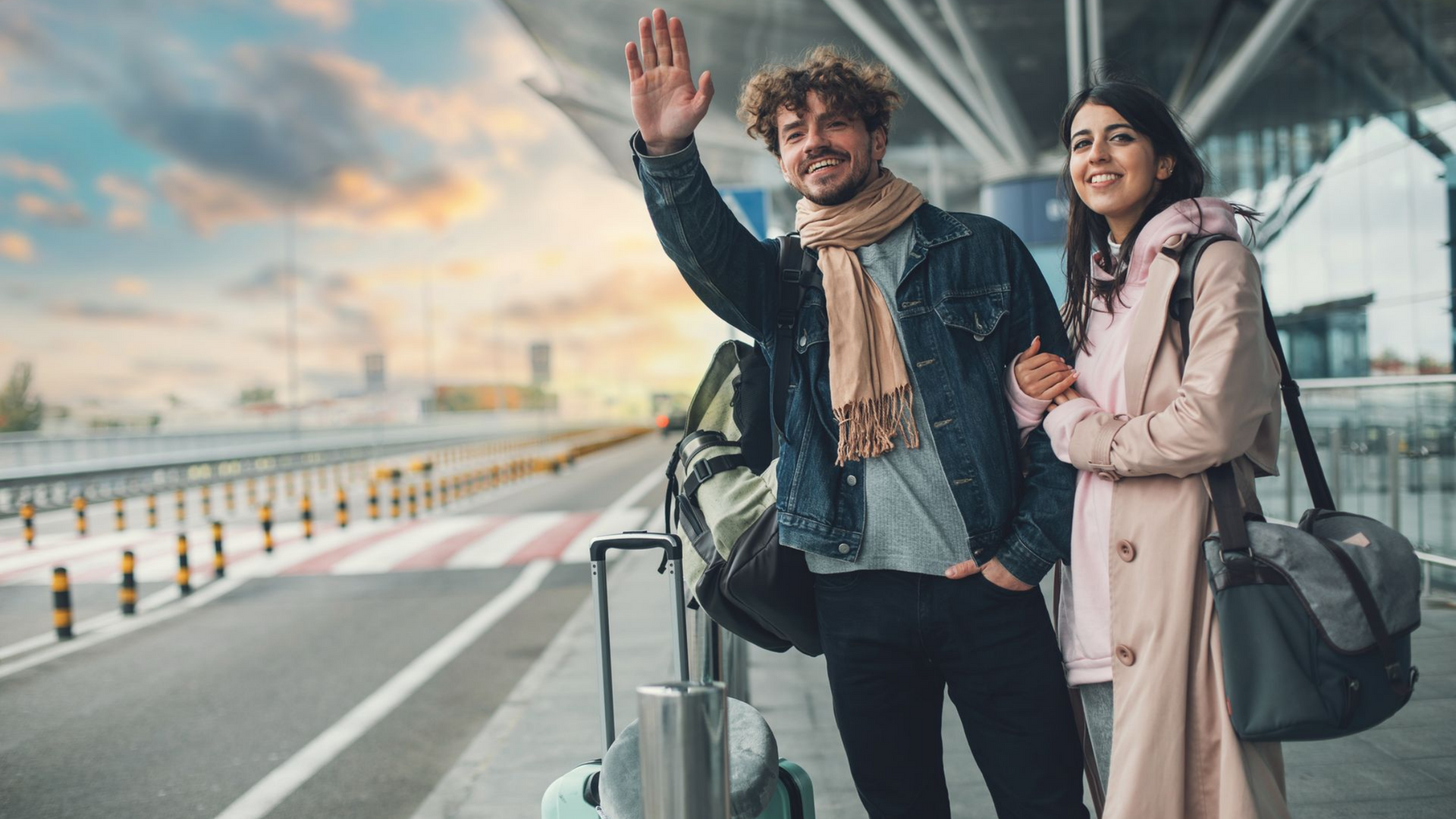 Un couple à l'aéroport ; l'homme fait signe de la main, la femme sourit, leurs bagages sont à proximité. Ciel nuageux.