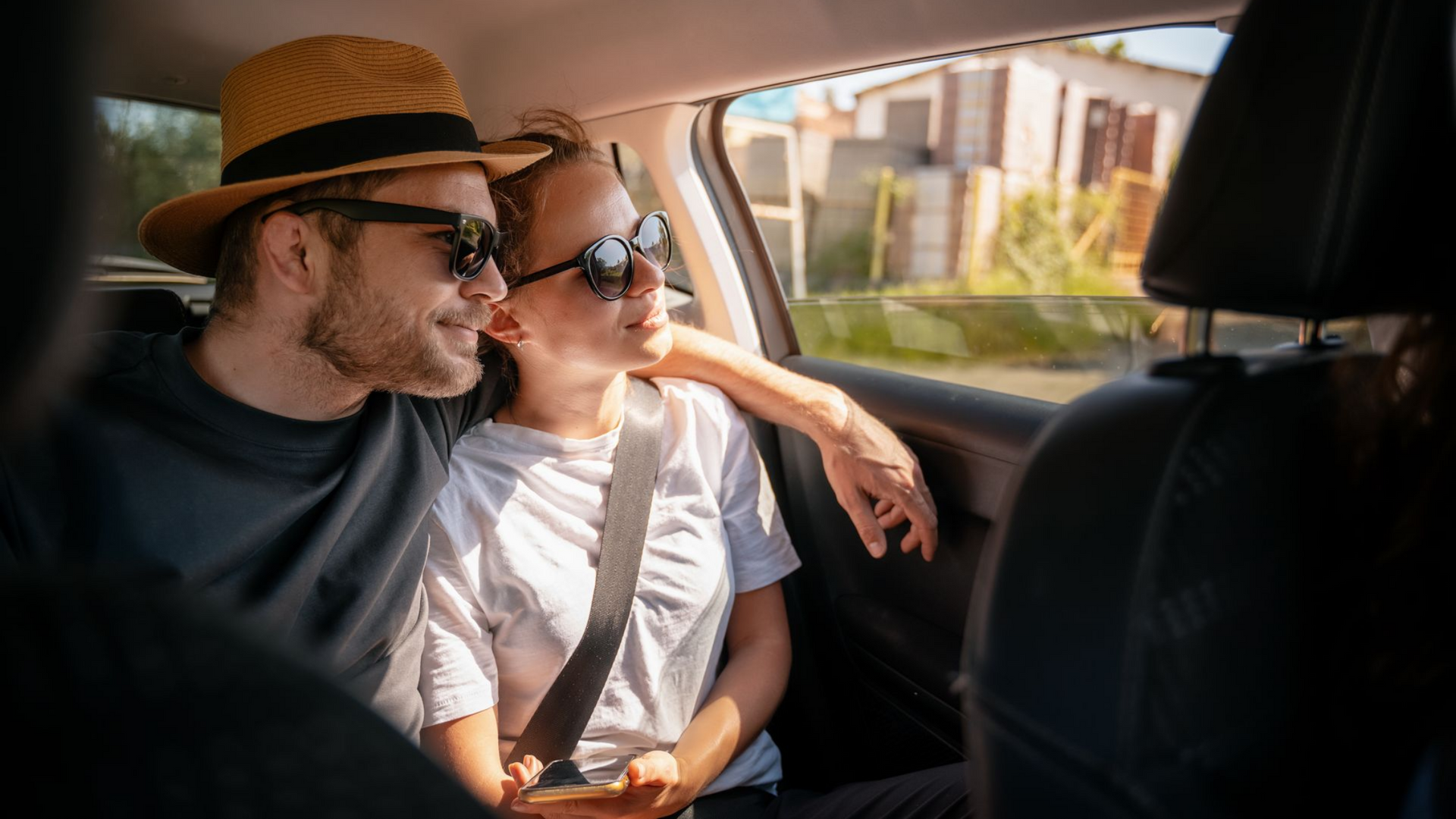 Un couple portant des lunettes de soleil, à l'intérieur d'une voiture, regardant par la fenêtre, le bras de l'homme autour de l'épaule de la femme.