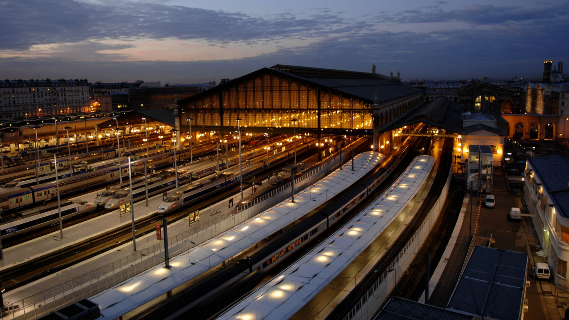 Vue nocturne d'une gare avec plusieurs voies, des bâtiments éclairés et des trains.