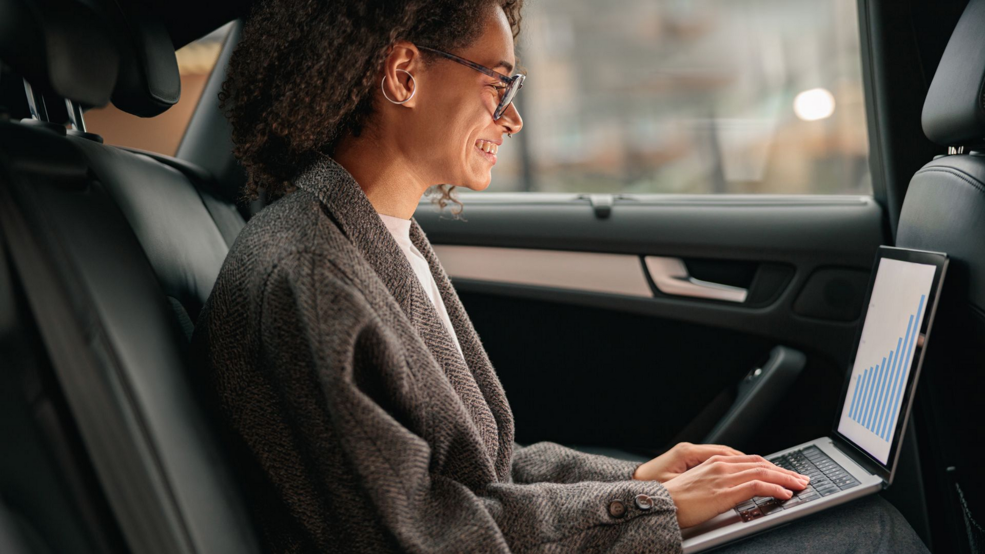 Femme en voiture, travaillant sur un ordinateur portable, souriante. Blazer gris, lunettes, intérieur en cuir noir.