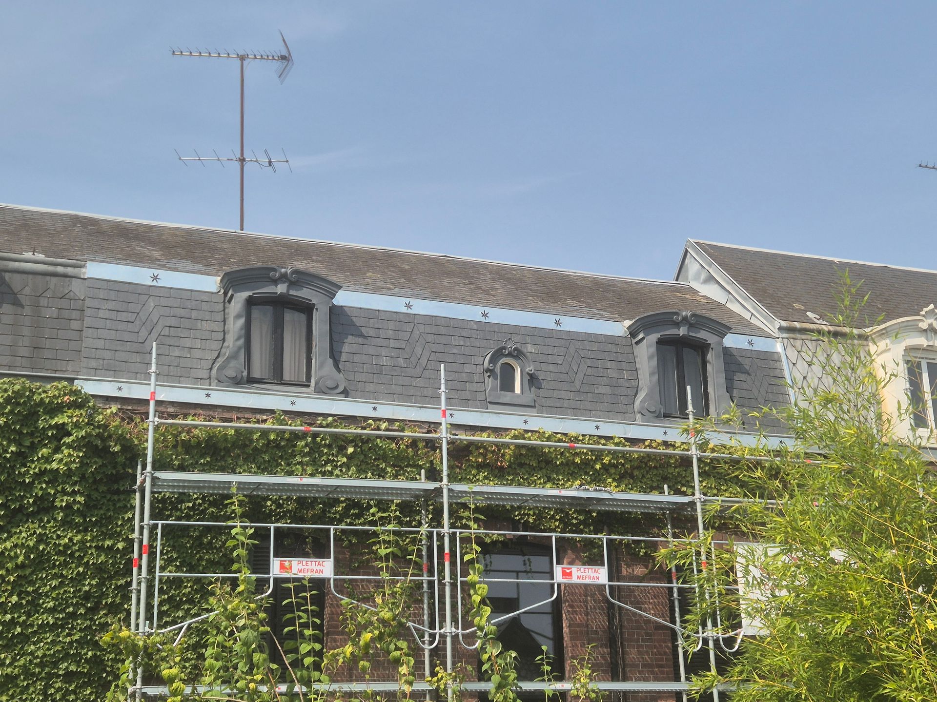 Bâtiment recouvert d'ardoise grise avec trois fenêtres à cadre blanc, chacune avec des jardinières remplies de fleurs rouges et vertes.