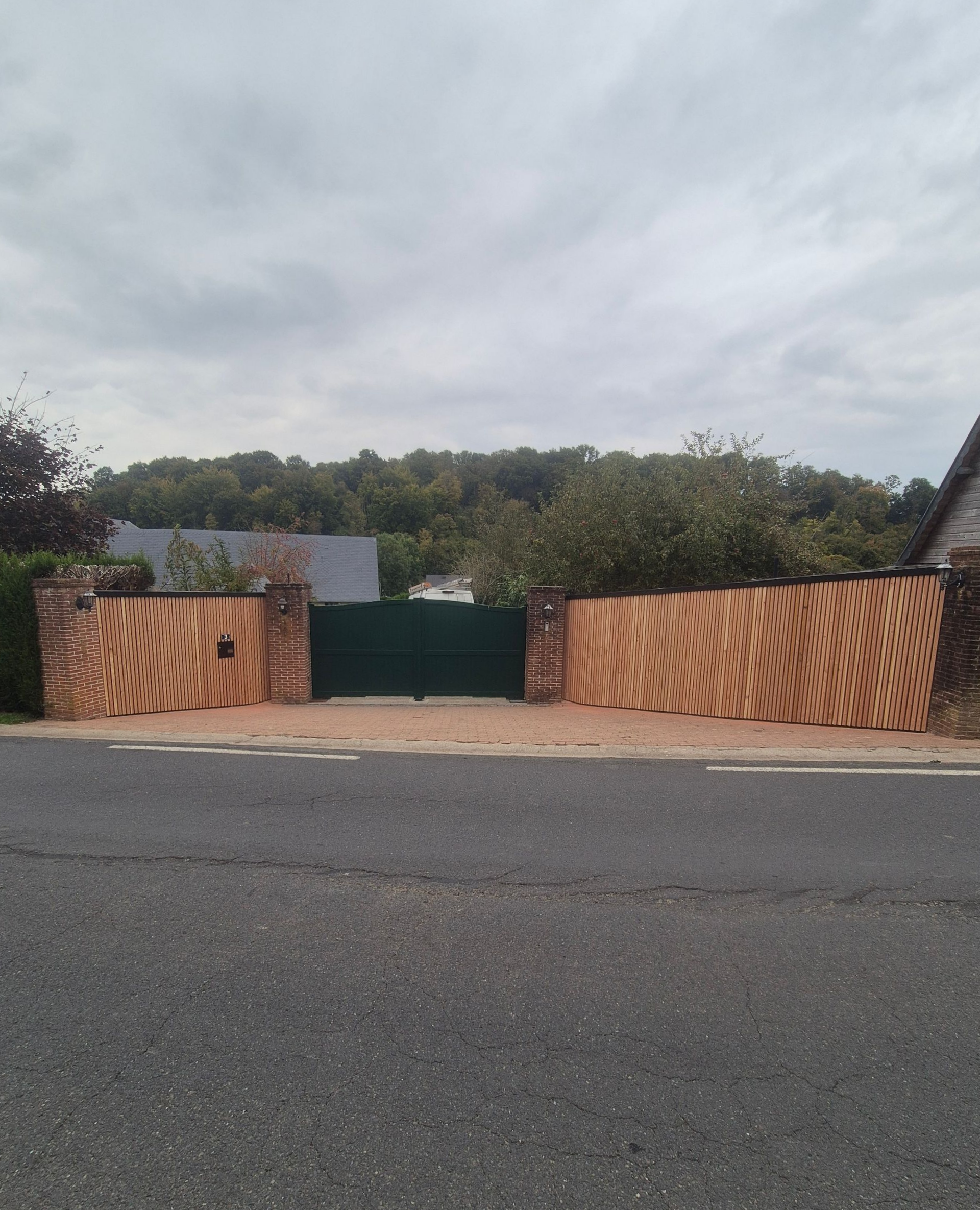 Façade en bois avec fenêtres à cadre noir, toit marron et luminaire extérieur. Arbres et ciel nuageux.