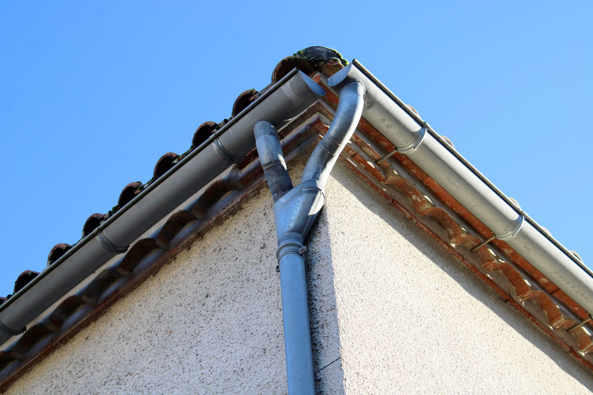 Coin d'une maison avec des gouttières grises, un tuyau de descente et un toit en tuiles contre un ciel bleu.
