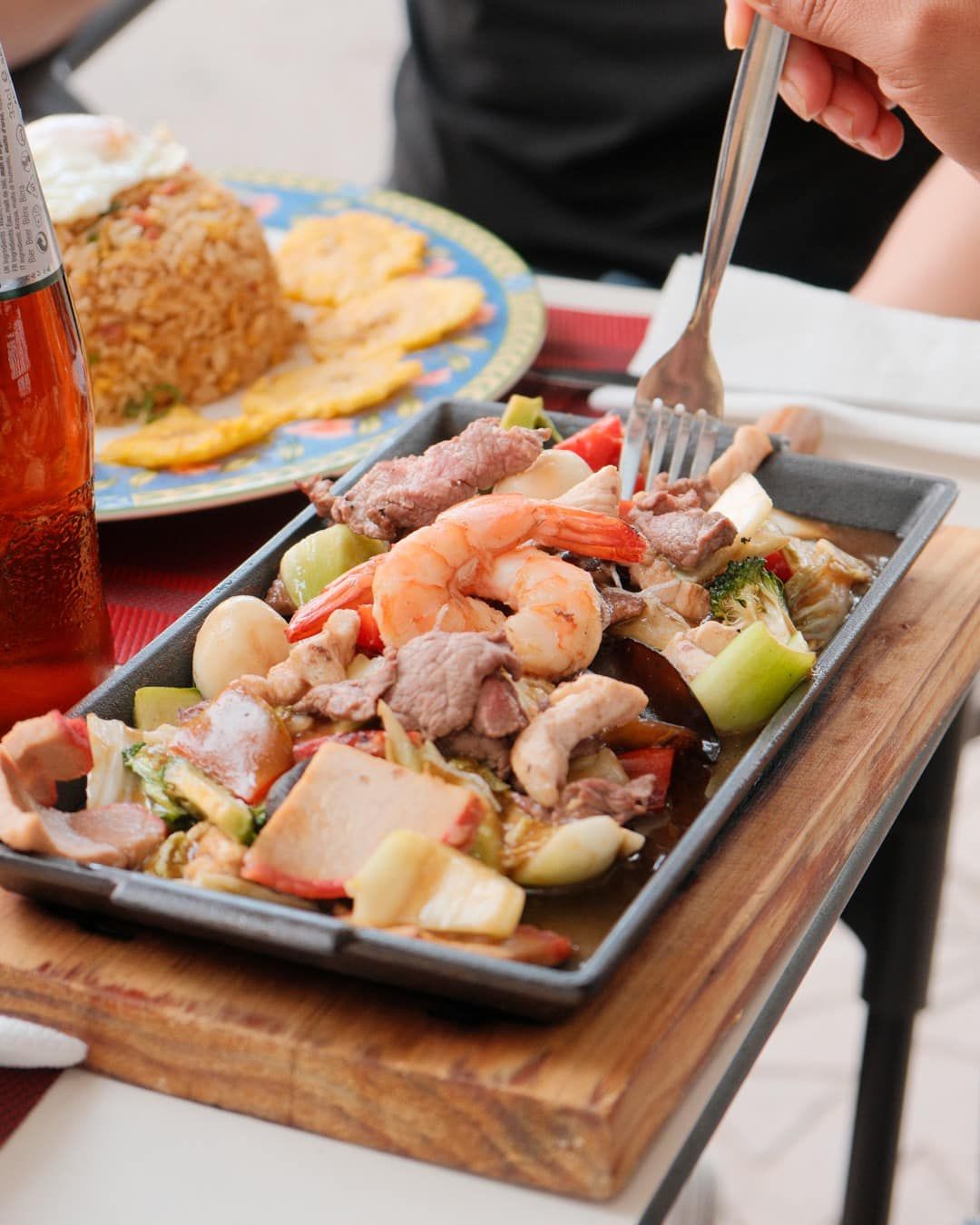 Plato de mariscos y carne con verduras, arroz frito y plátanos en una mesa. Una mano sostiene un tenedor.