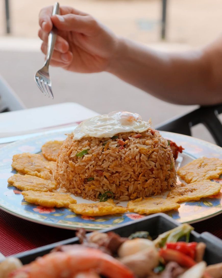 Plato de arroz frito con huevo frito, tostones y una mano sosteniendo un tenedor, en un entorno al aire libre.