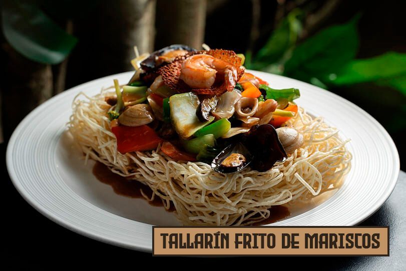 Fideos fritos con mariscos en un plato blanco con verduras y salsa. El texto dice 