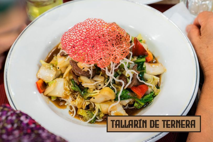 Plato de fideos con carne de res, bok choy, pimiento rojo y una crujiente decoración, servido en un tazón blanco.