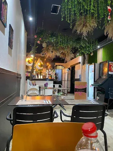 Interior de restaurante con mesas y sillas. Decoración con plantas colgantes y techo negro.