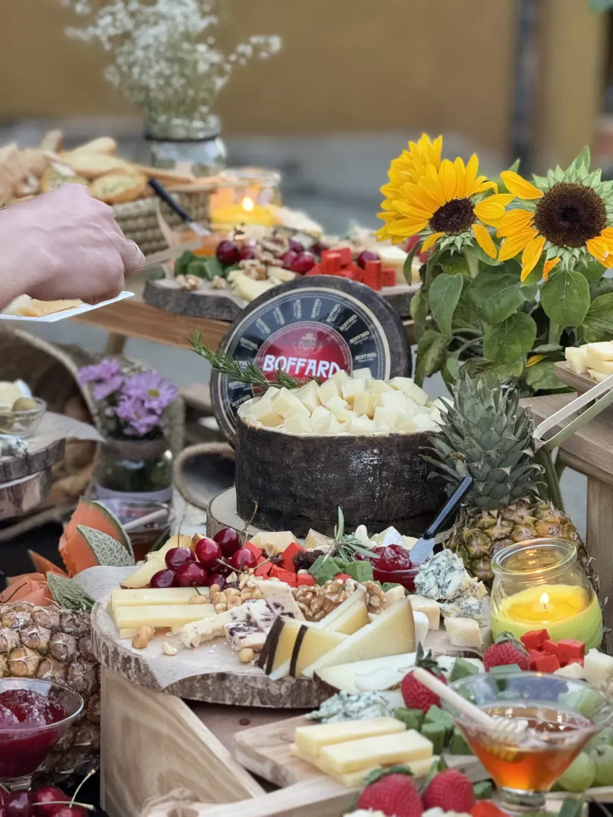 Tabla de embutidos con diversos quesos, frutas y nueces, con girasoles y una persona tomando comida.