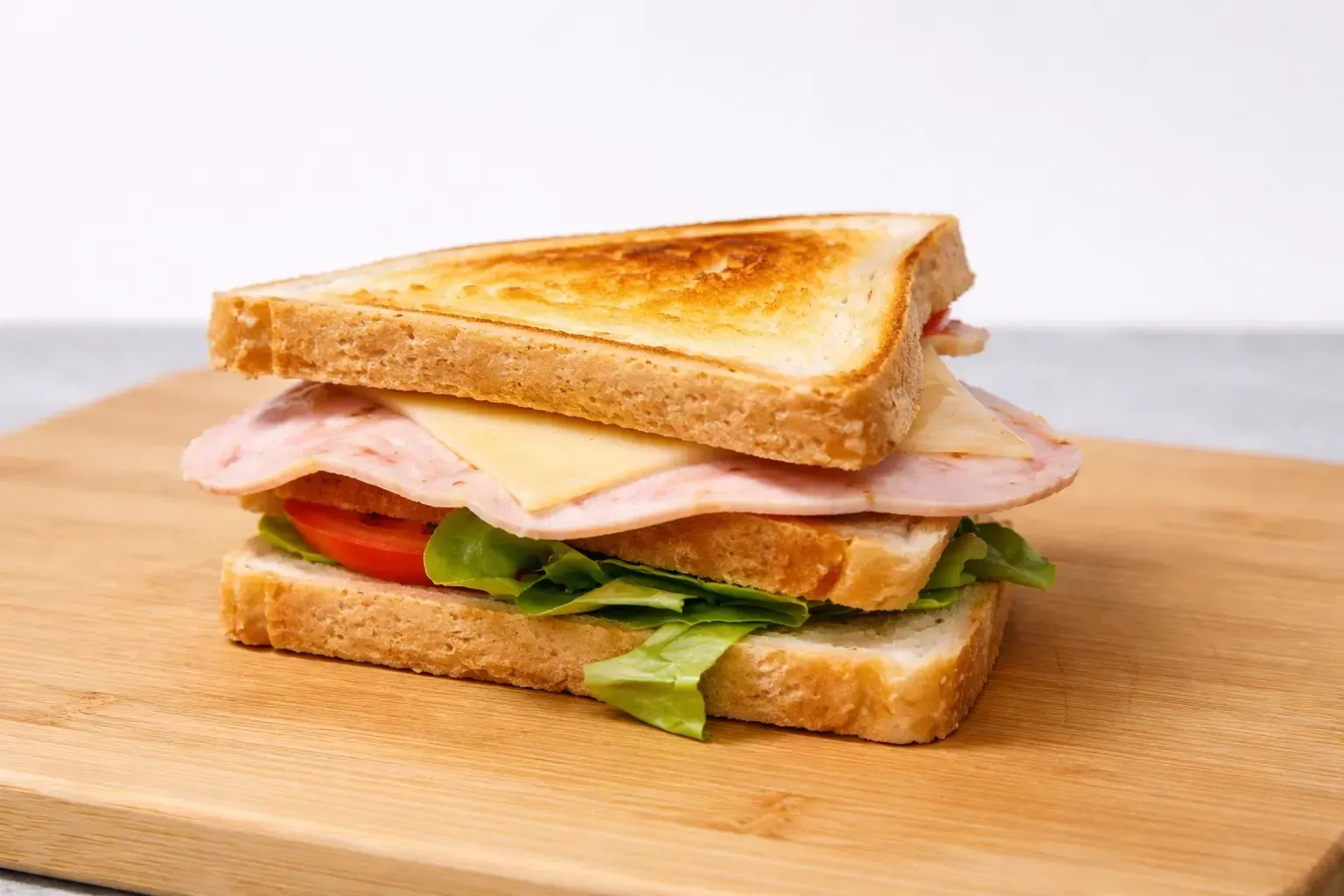 Sándwich sobre tabla de madera: pan tostado, capas de jamón, queso, tomate y lechuga.