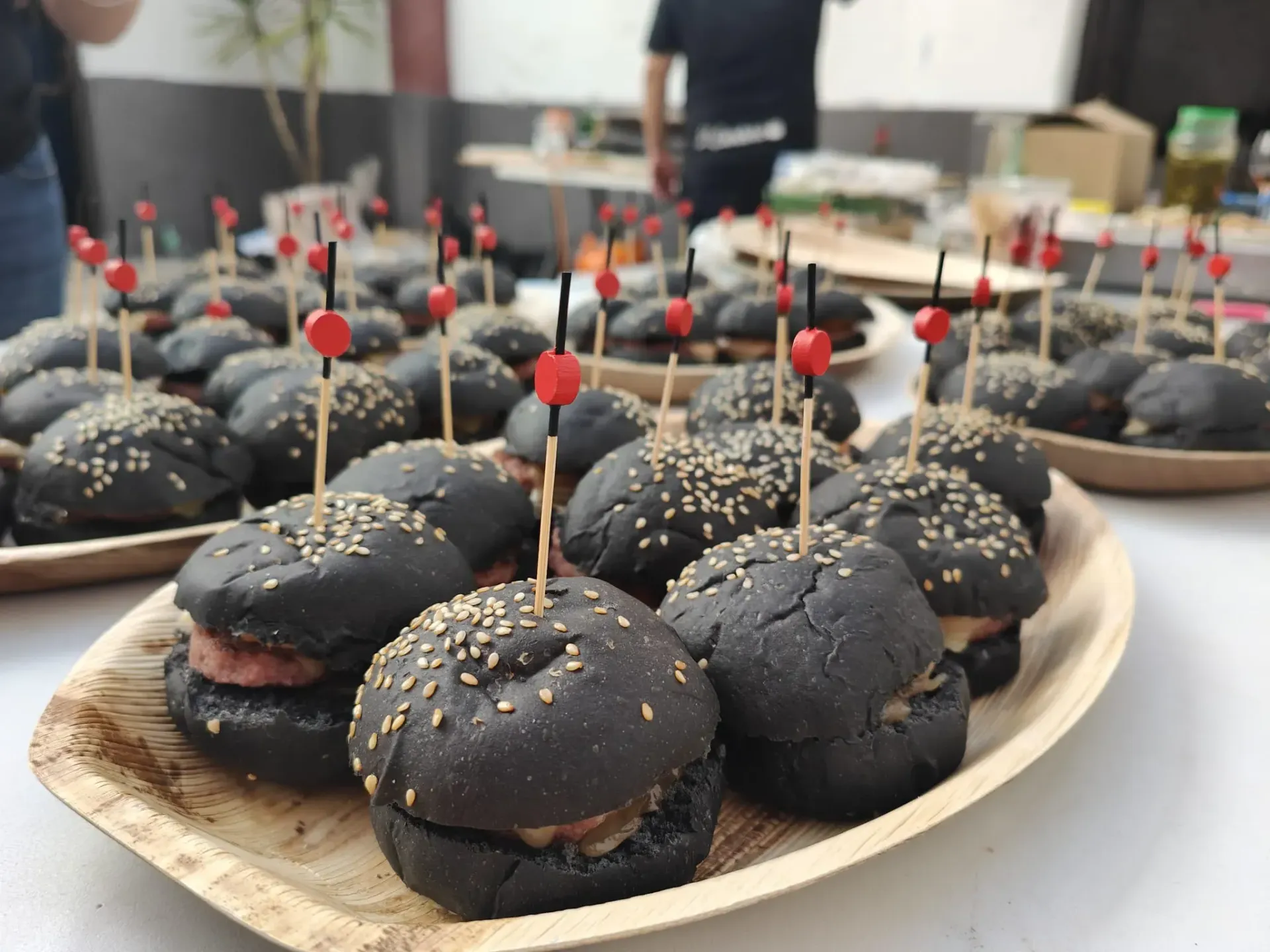 Minihamburguesas negras en platos con forma de hoja, cada una de ellas ensartada con un palillo con punta roja.