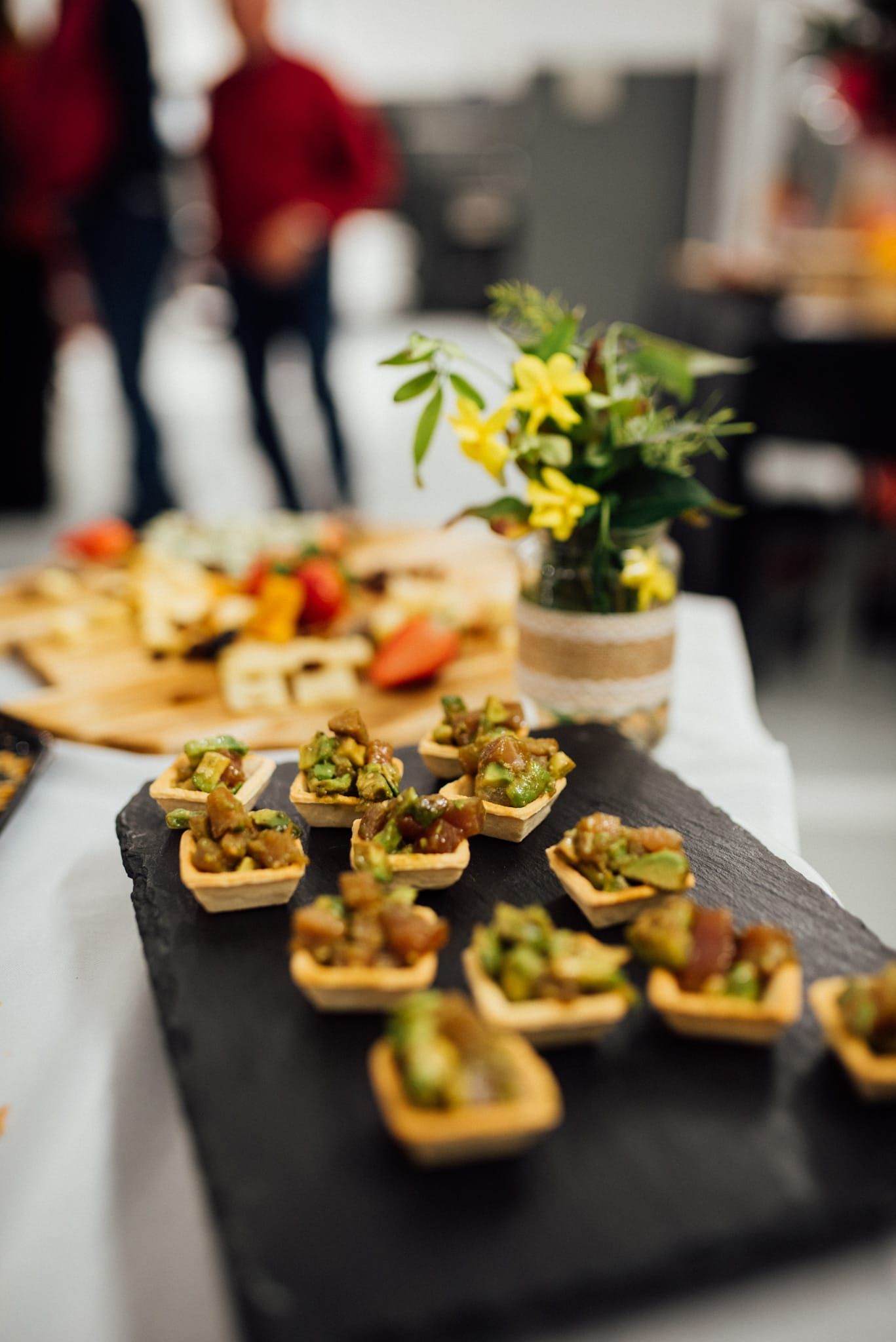 Aperitivos en una pizarra, una tabla de quesos y un pequeño arreglo floral sobre una mesa. Dos personas al fondo.