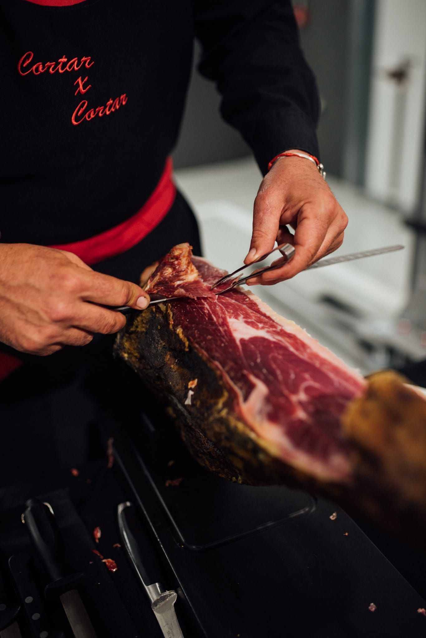 Persona cortando jamón con un cuchillo largo, vestida con camisa negra y delantal rojo.