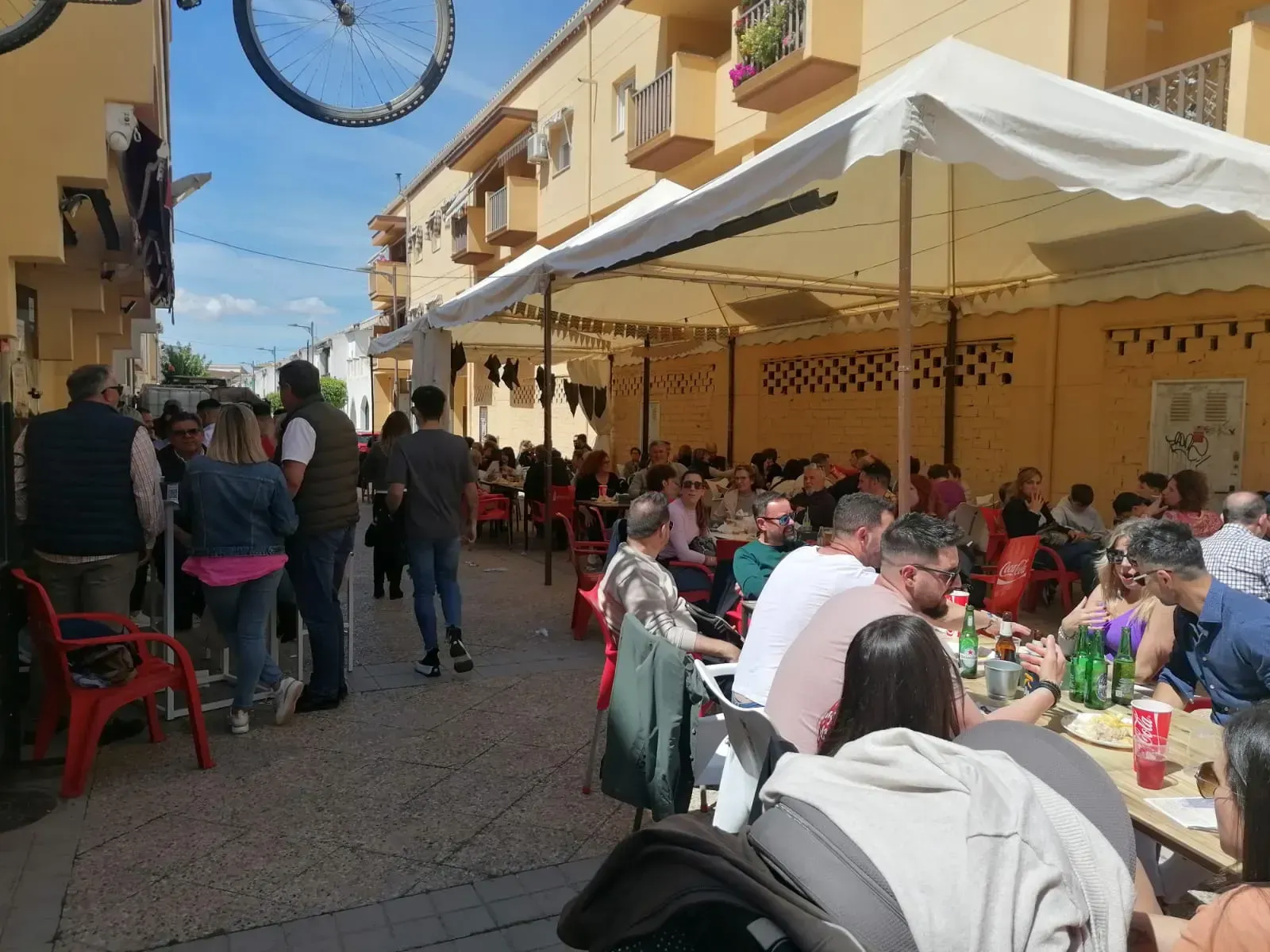 La gente se reunió en mesas al aire libre bajo toldos blancos, en una calle con edificios amarillos.