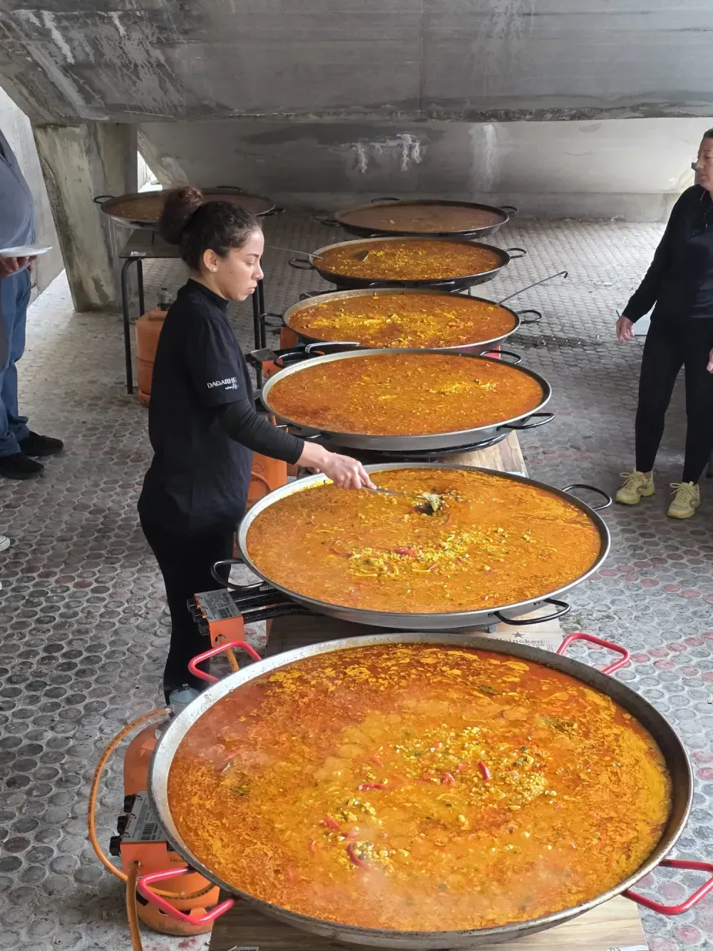 Una mujer revuelve la paella en grandes sartenes al aire libre, mientras varias otras sartenes están listas.