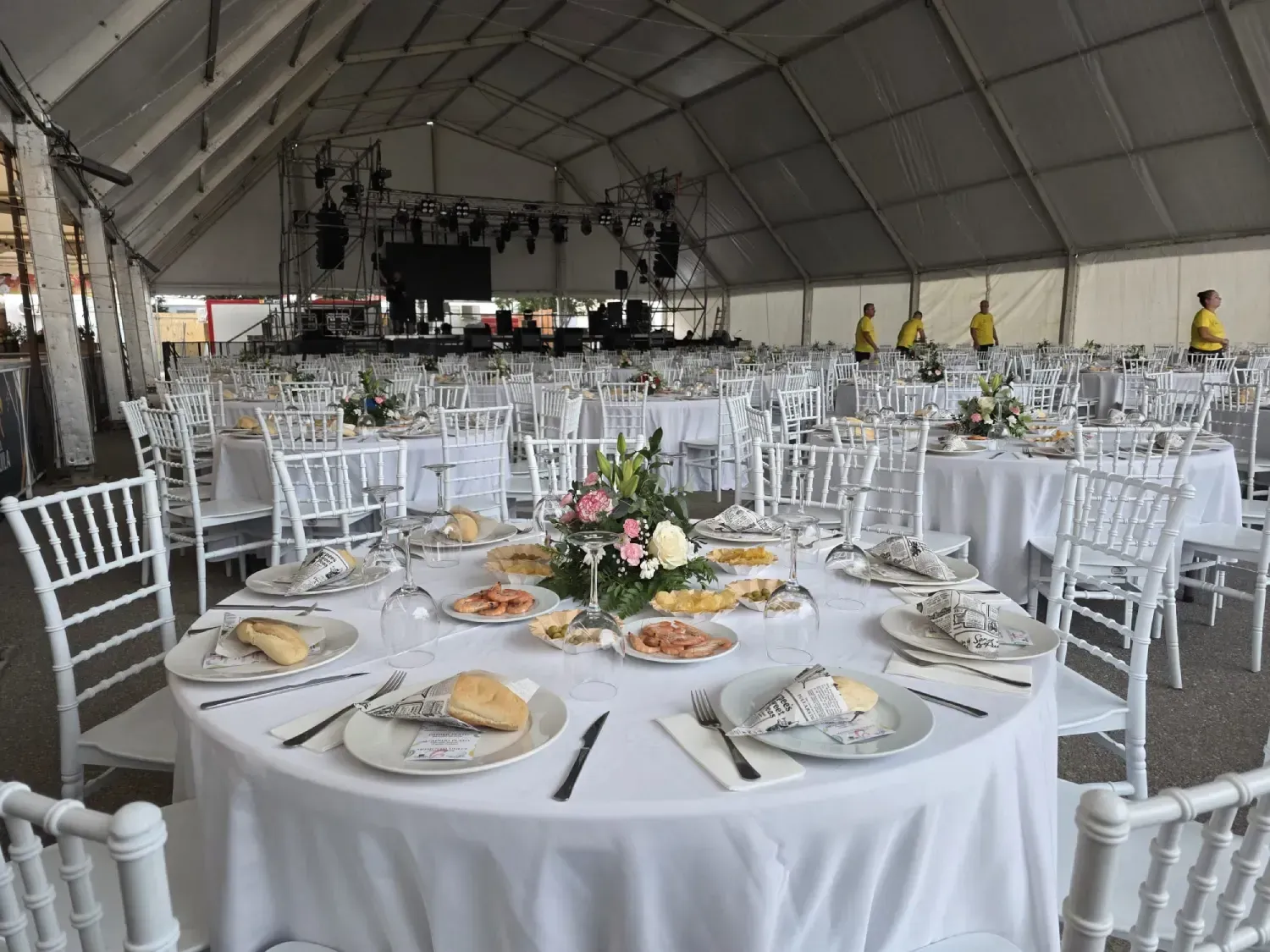 Mesas preparadas para un evento dentro de una gran carpa blanca, escenario visible al fondo.