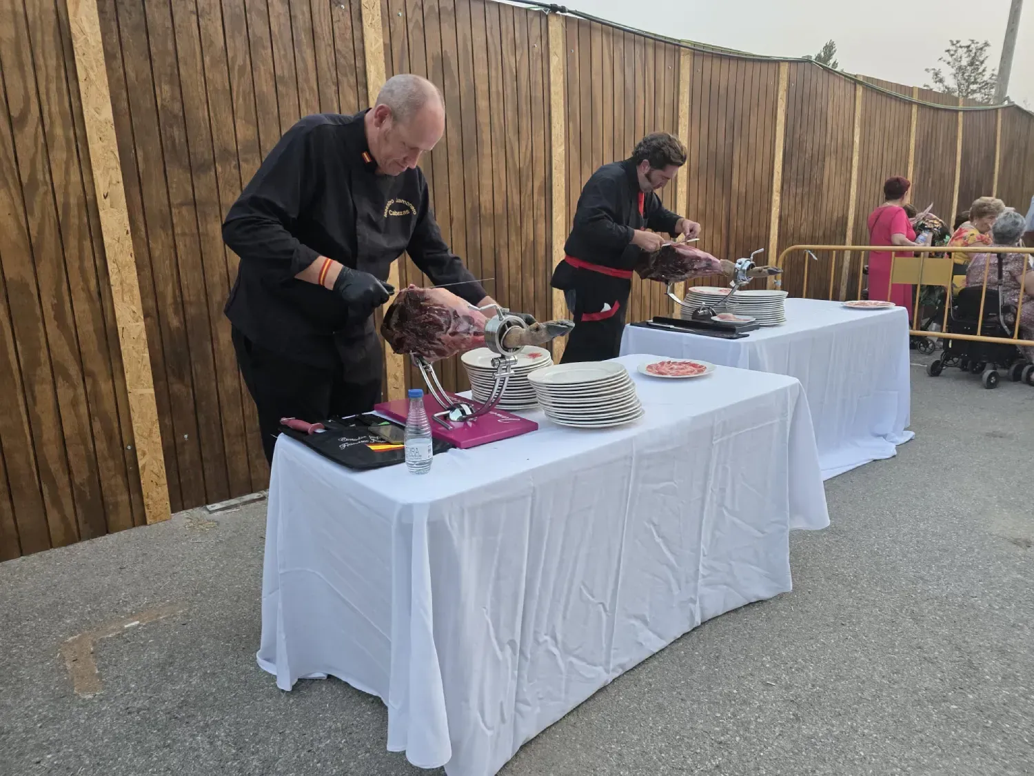 Dos cocineros cortan jamón en mesas blancas al aire libre; hay una valla de madera al fondo.