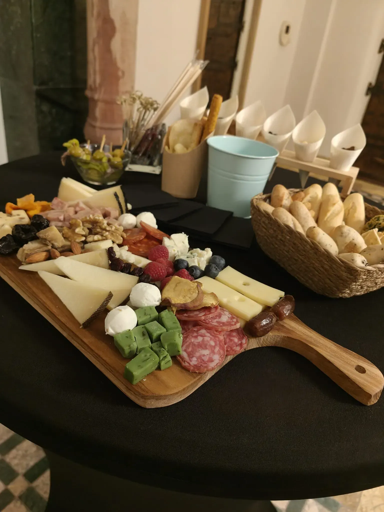 Tabla de quesos y embutidos con diversos aperitivos sobre una mesa negra. También hay pan, aceitunas y palitos de pan.