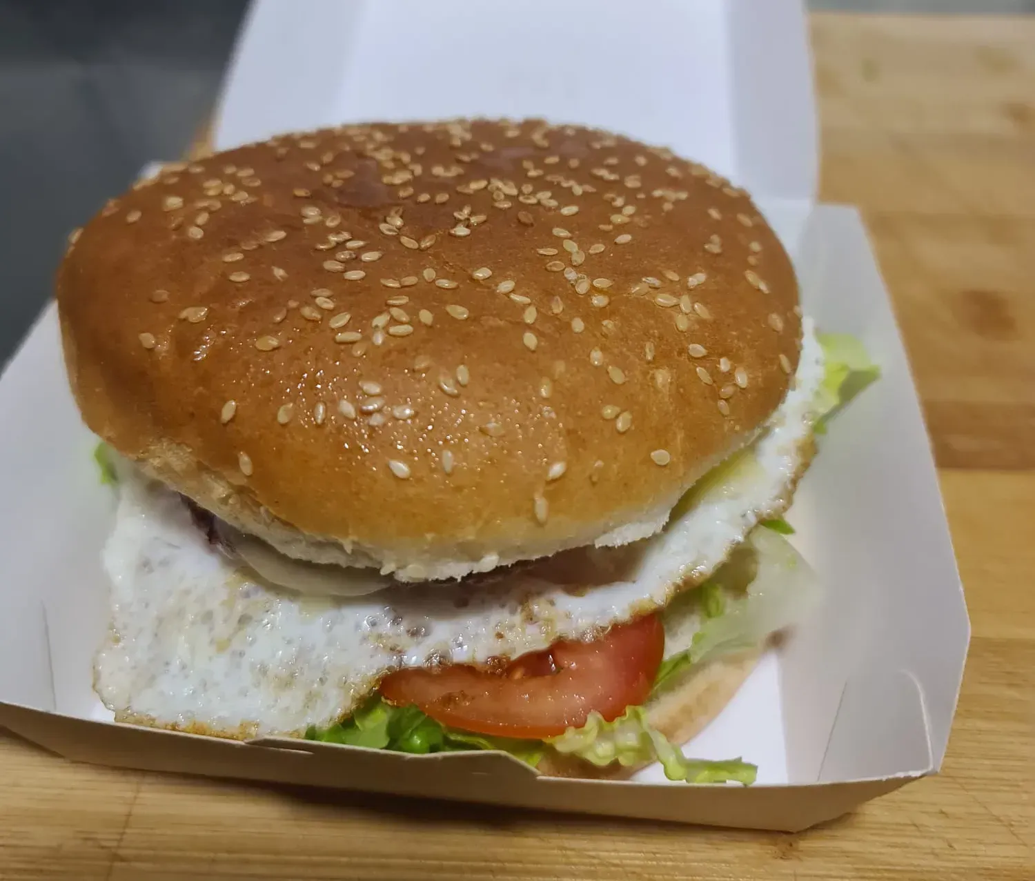 Hamburguesa en caja blanca con pan de semillas de sésamo, lechuga, tomate y huevo frito.