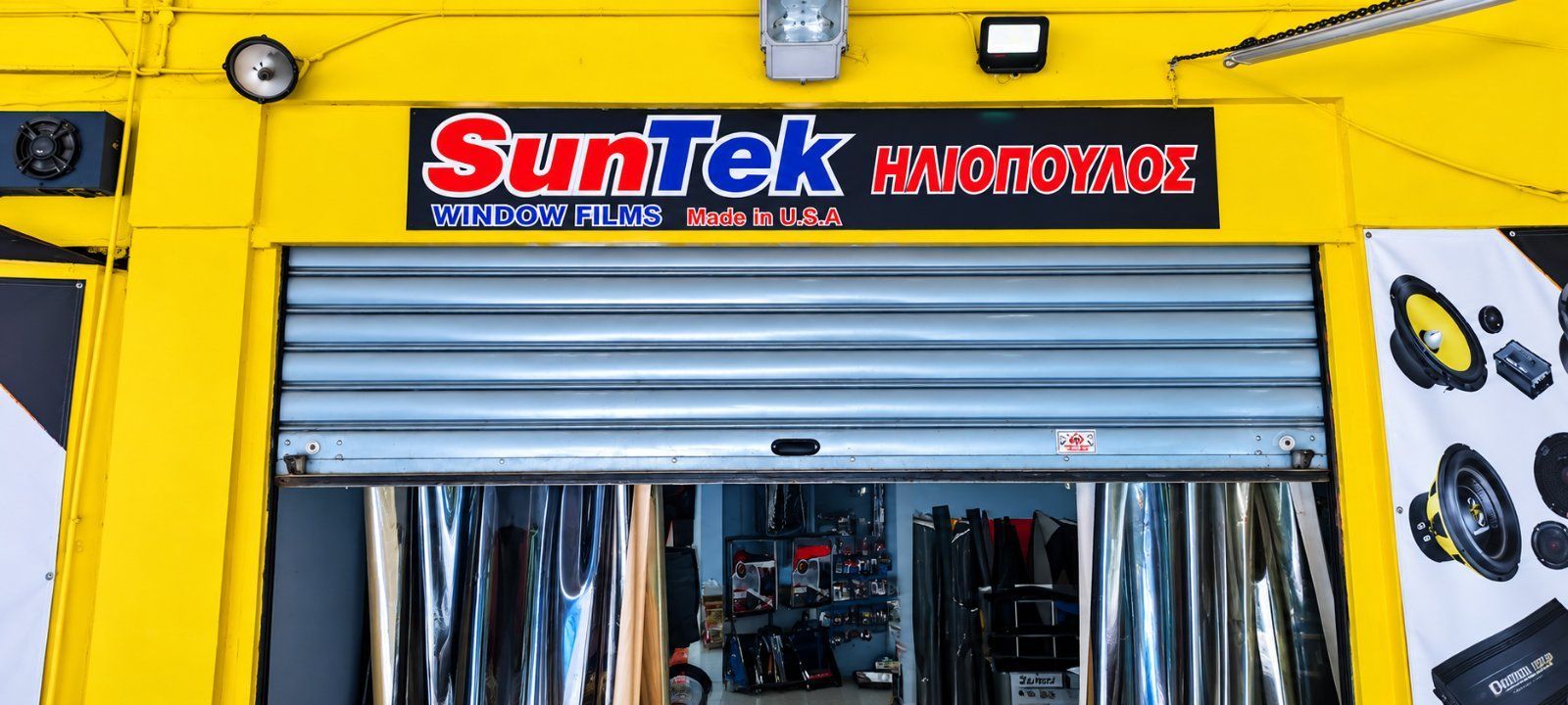 Κίτρινη πρόσοψη καταστήματος με ασημί ρολό ασφαλείας και επιγραφή SunTek πάνω από την είσοδο.