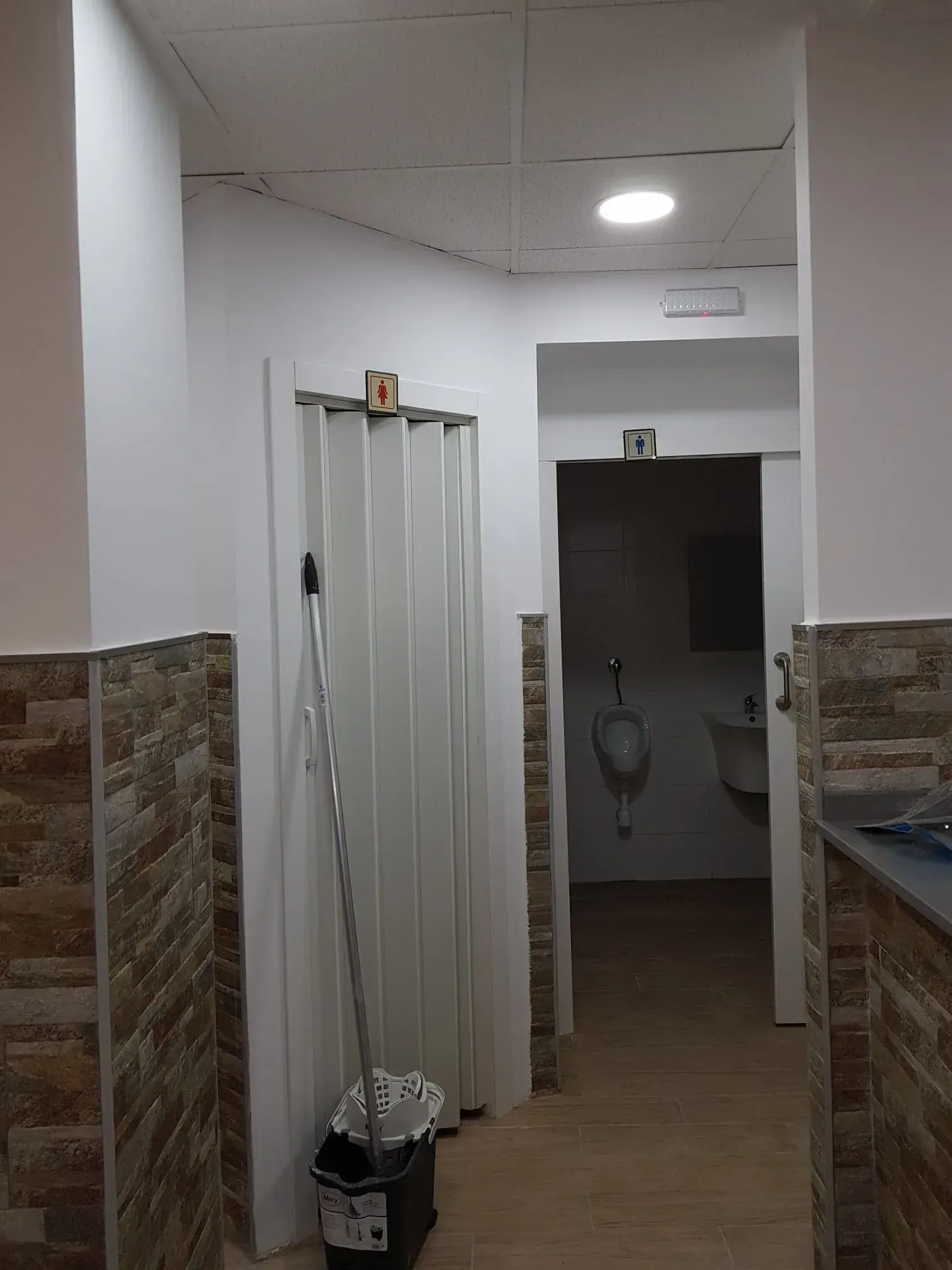 Pasillo estrecho con paredes alicatadas, un marco de puerta y un baño poco iluminado visible al final.