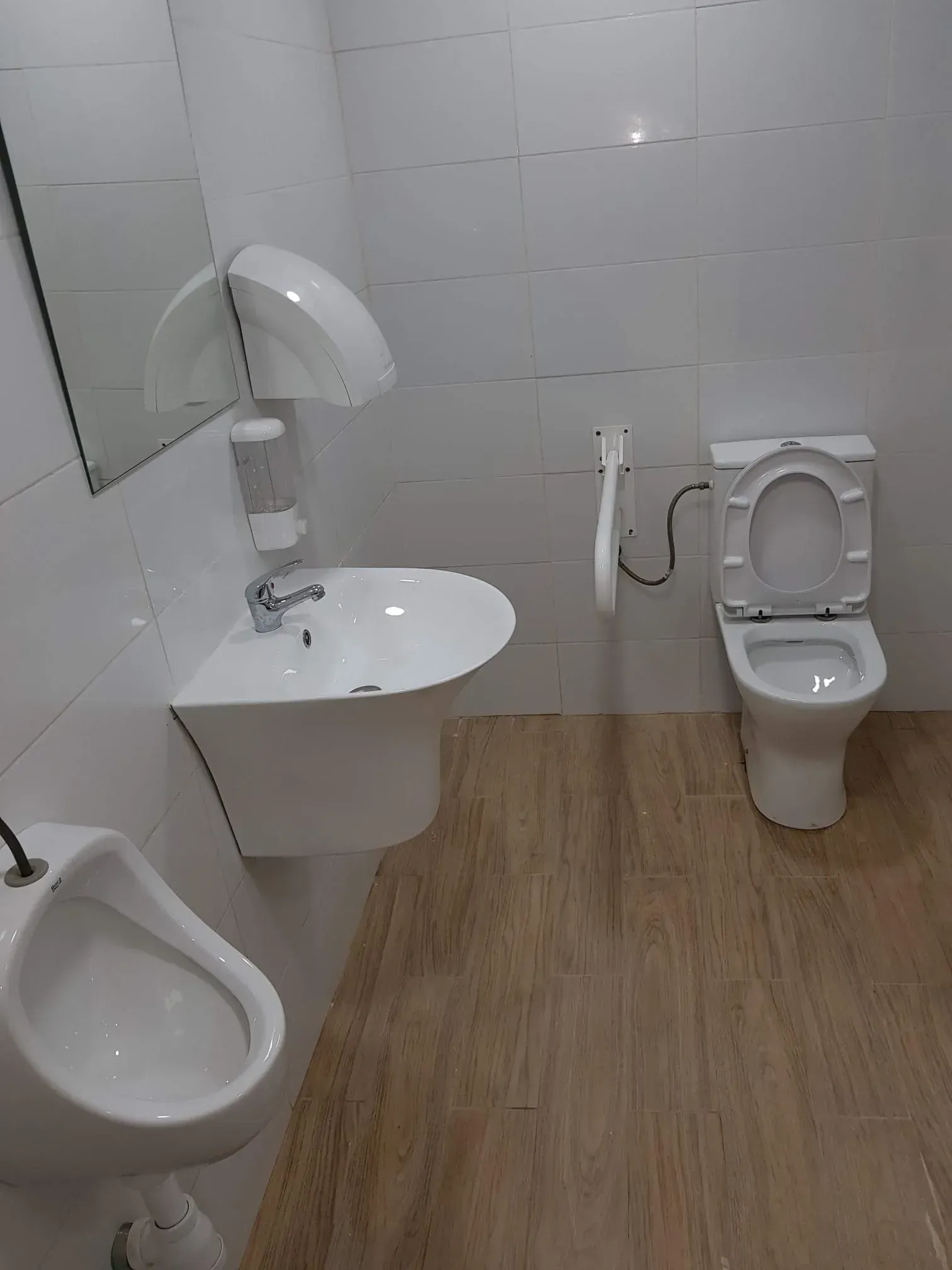 Baño accesible con lavabo, inodoro y urinario mural sobre paredes alicatadas en blanco y suelo con estampado de madera.