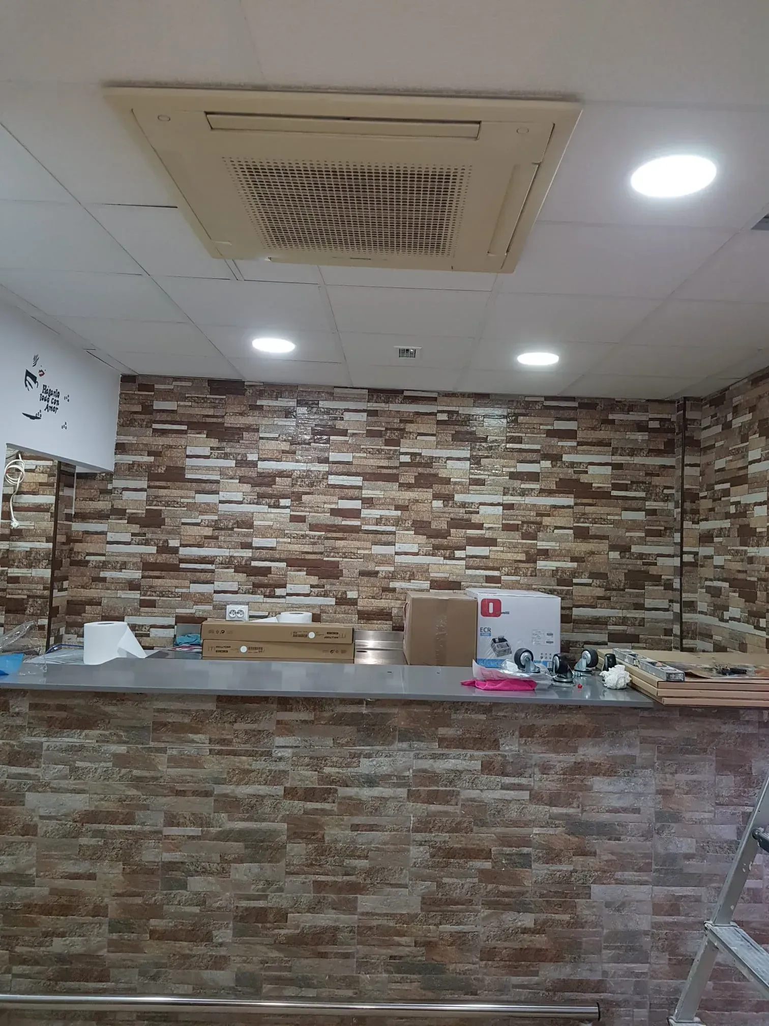 Mostrador interior con suministros apilados y una pared con diseño de piedra, bajo luces de techo y una rejilla de ventilación.