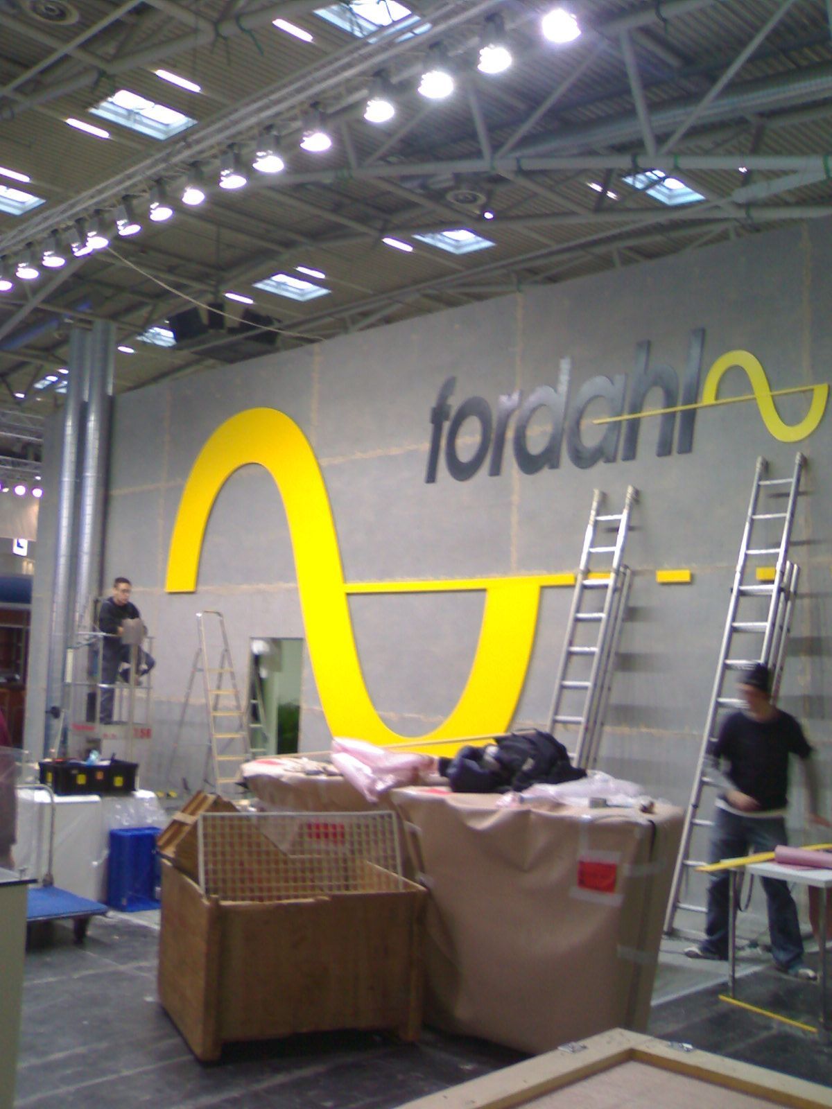 Arbeiter montieren ein gelbes „Fordahl“-Logo an einer grauen Wand. Leitern und Ausrüstung sind vorhanden.