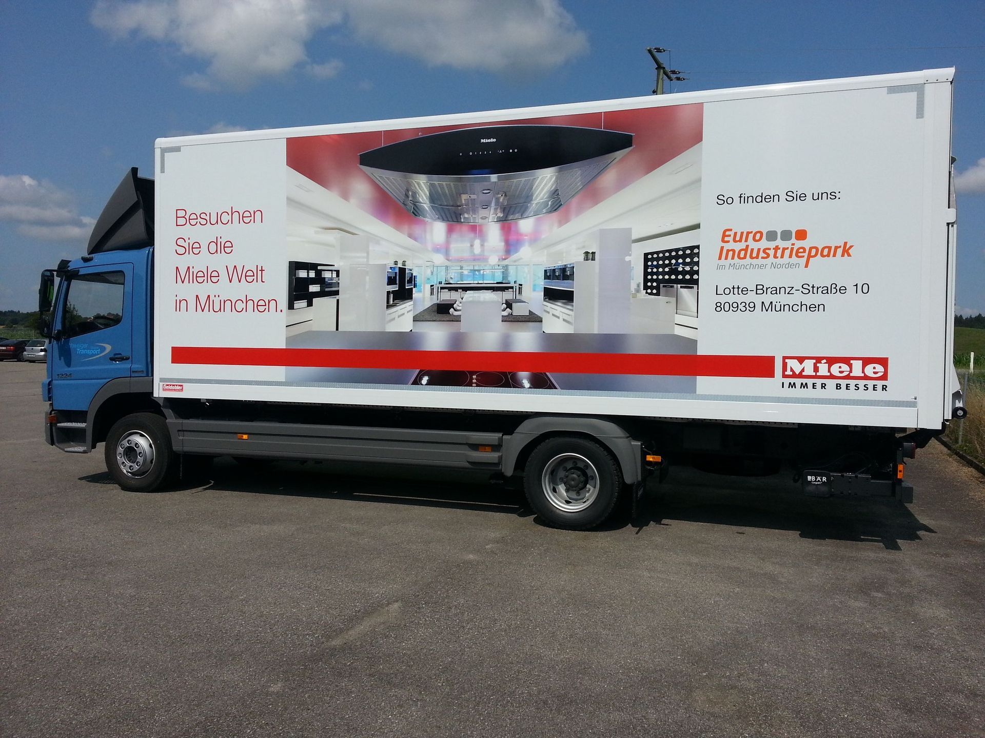 Blauer Miele-LKW mit weißer Werbung, geparkt auf dem Bürgersteig vor einem sonnigen, blauen Himmel.