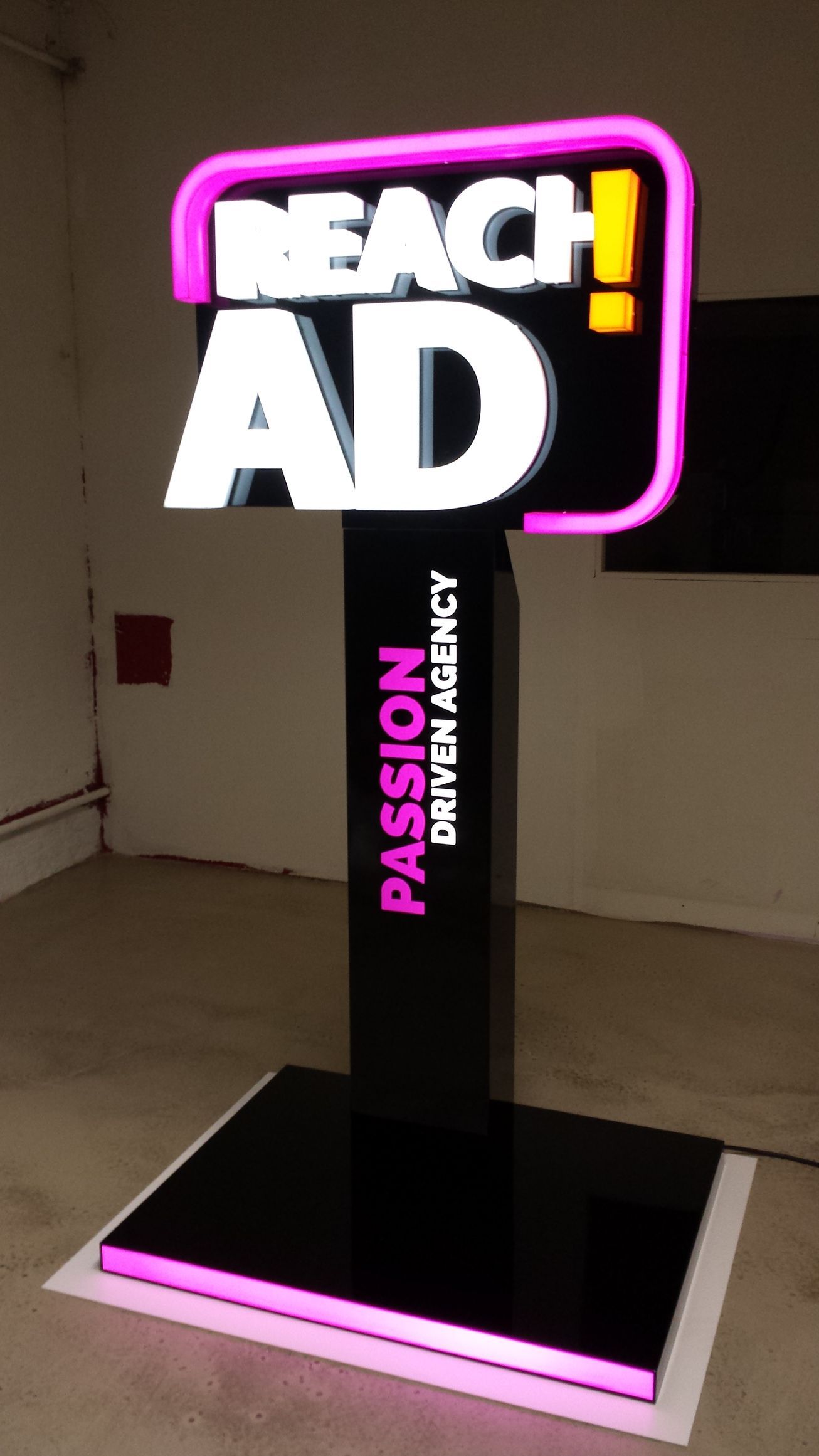 Schild für „Reach Ad“ mit neonpinker und weißer Schrift auf schwarzem Ständer. „Passion Driven Agency“ ist in Pink.