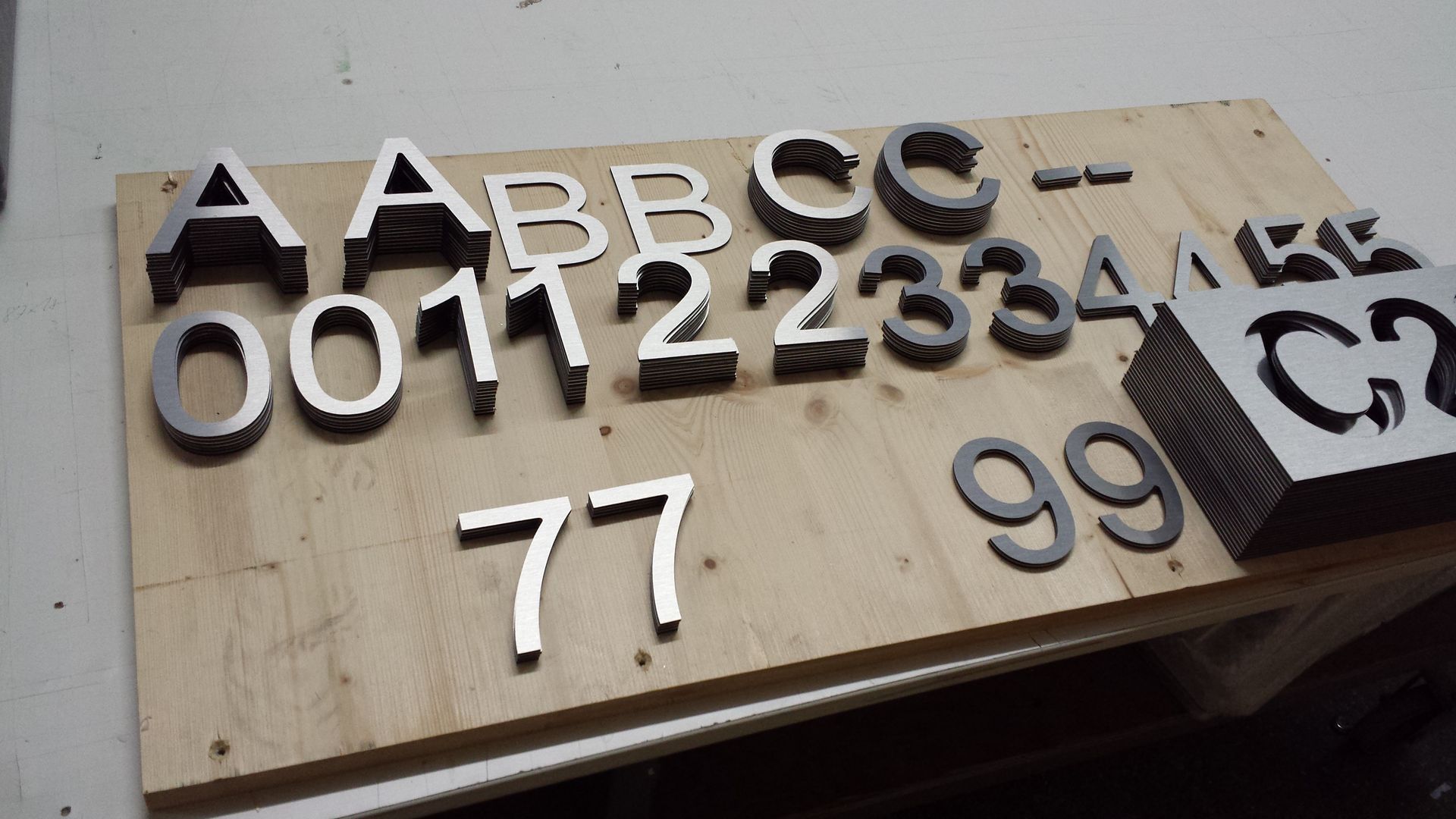 Auf einer Holzoberfläche angeordnete Metallbuchstaben und -zahlen, darunter A, B, C, 0, 1, 2, 3, 4, 7 und 9.