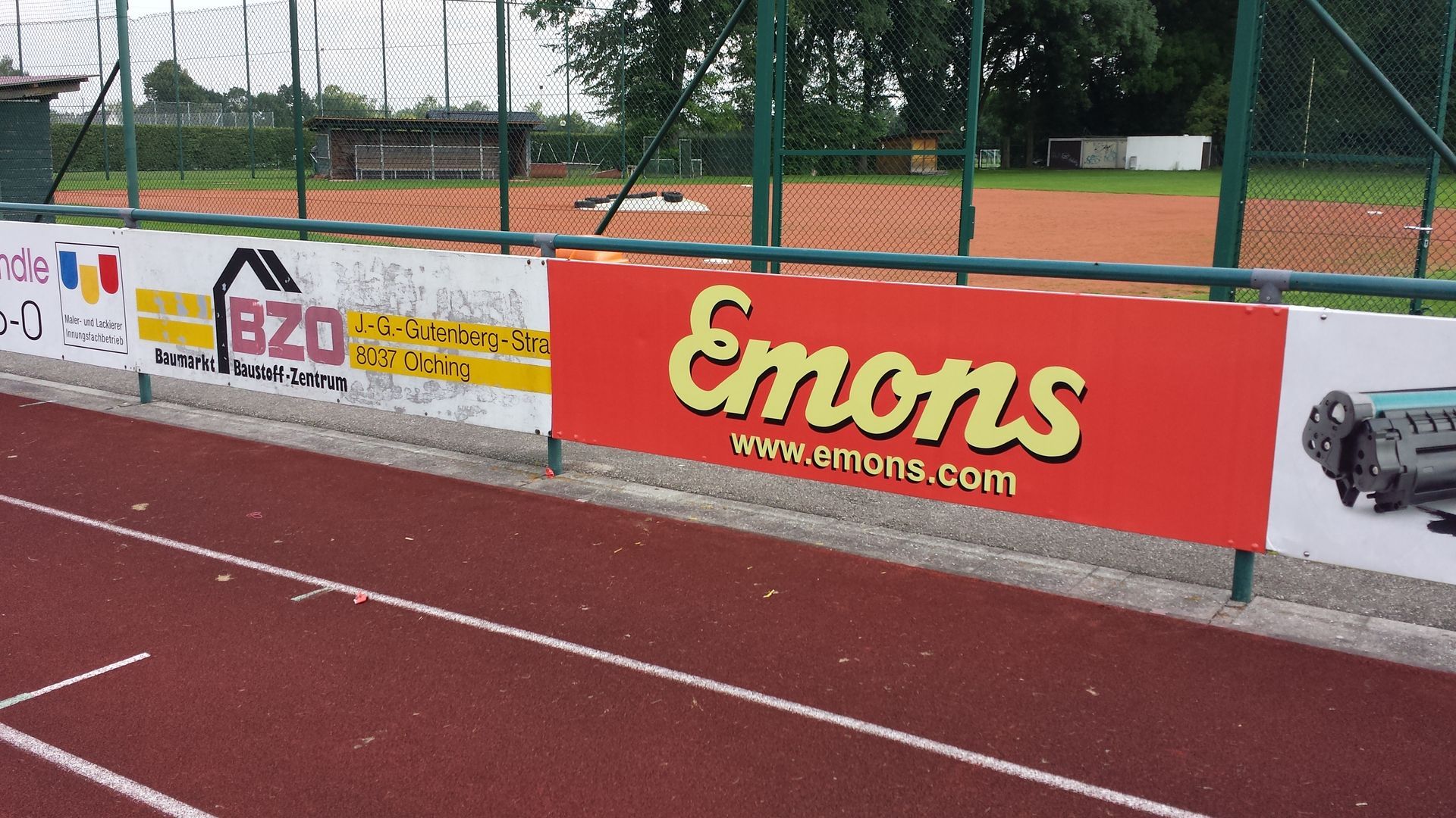 Sportplatz mit Bandenwerbung, „Emons“ in Gelb auf Rot.