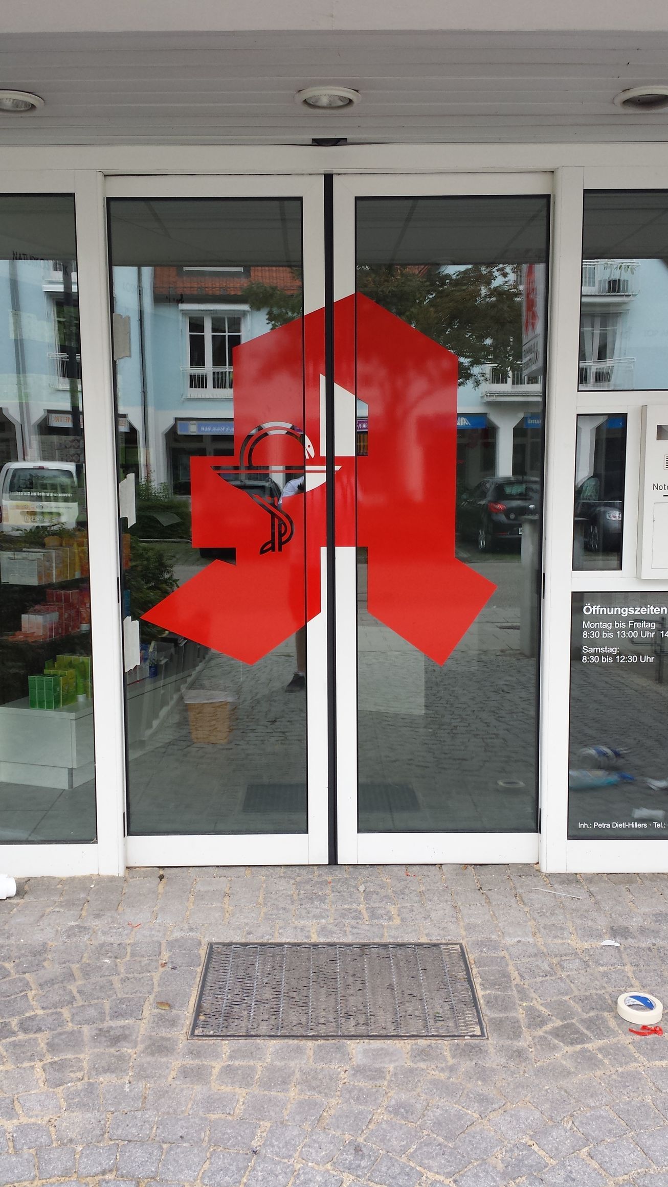 Rotes Apothekensymbol auf Glastüren. Eingang zur Apotheke über einen gemauerten Weg.