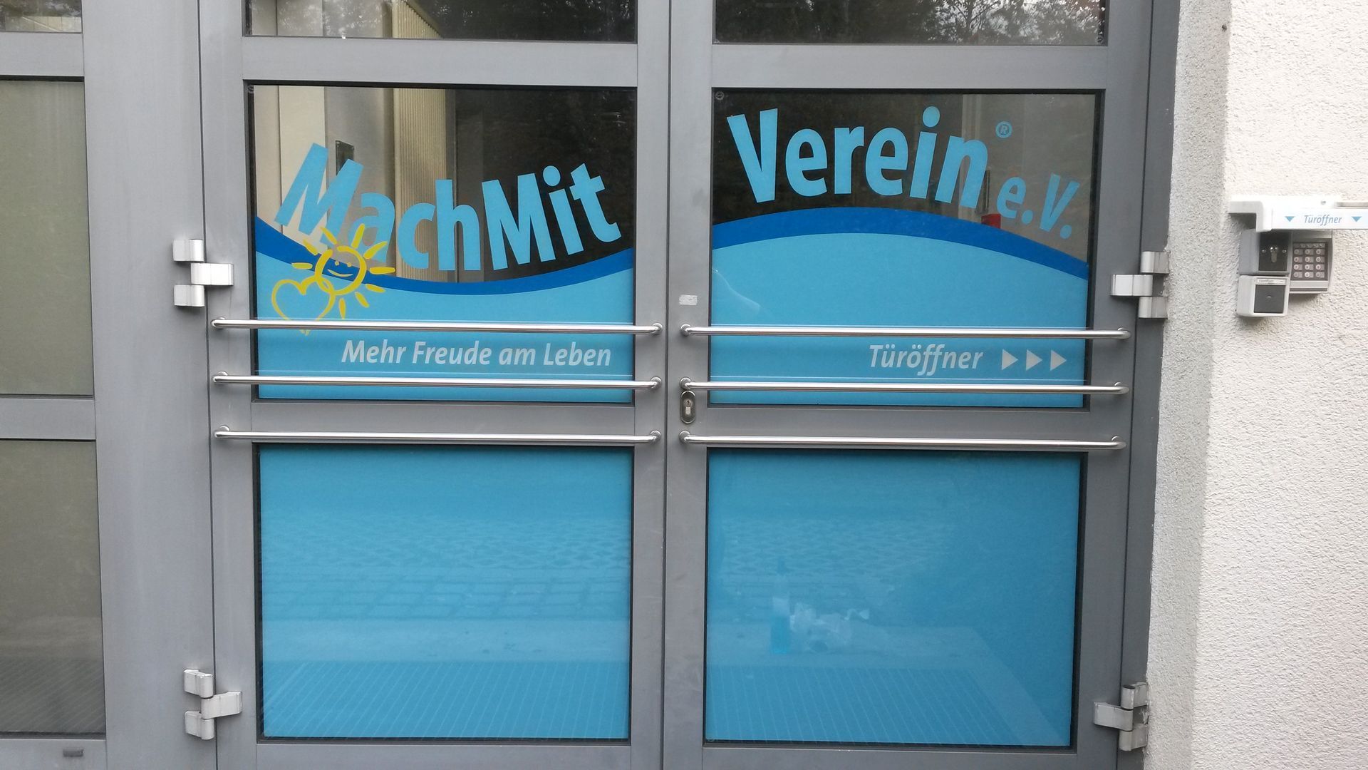 Doppeltür mit „MachMit Verein eV“-Logo, blauem Wellendesign und Text an einer Gebäudeaußenseite.